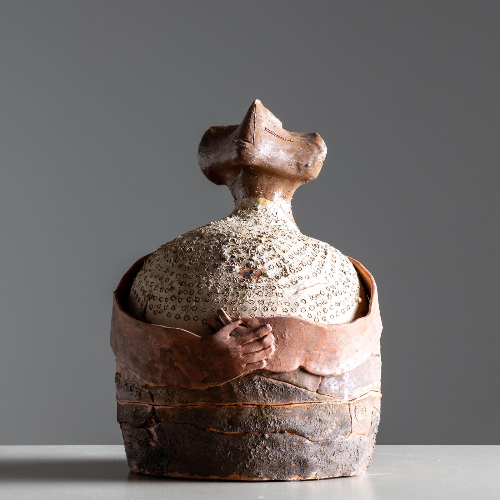 Scultura antropomorfa in terracotta con corpo bulboso e braccia lunghe. Le mani sono intrecciate e la testa a forma di cuneo ha occhi, naso e bocca. La superficie presenta diverse texture ed è parzialmente smaltata e patinata. La scultura è firmata