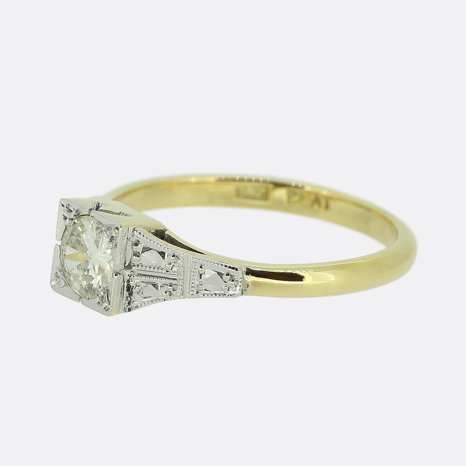 Hier haben wir einen diamantenen Verlobungsring aus 18 Karat Gelbgold. Der Diamant ist ein runder, facettierter Übergangsschliff und in einer quadratischen Platinfassung gefasst, die sein Funkeln zur Geltung bringt. Jede Schulter ist außerdem mit