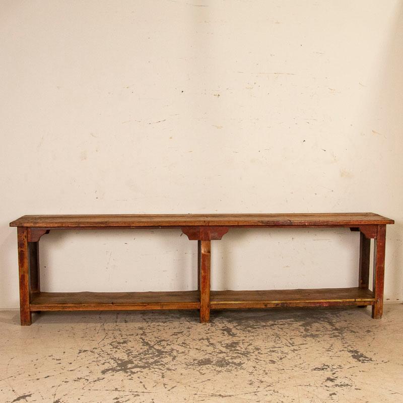 At just under 10’ long, it is the incredible aged patina that invites one to run your fingers down the long top of this striking console table. The natural pine top and shelf are complimented by the original painted brick red legs while even the