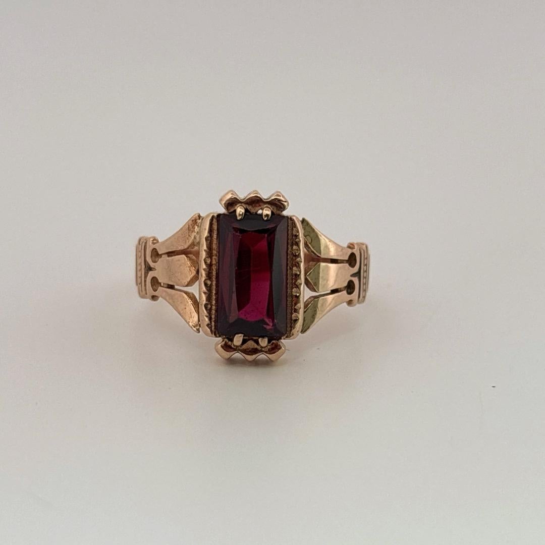 Dieser exquisite antike Ring ist aus warmem 10-karätigem Ros�égold gefertigt und zeigt einen auffälligen, tiefroten Granat als Herzstück. Der Granat in eleganter Smaragdform strahlt einen satten, weinähnlichen Farbton aus, der das Licht mit subtiler