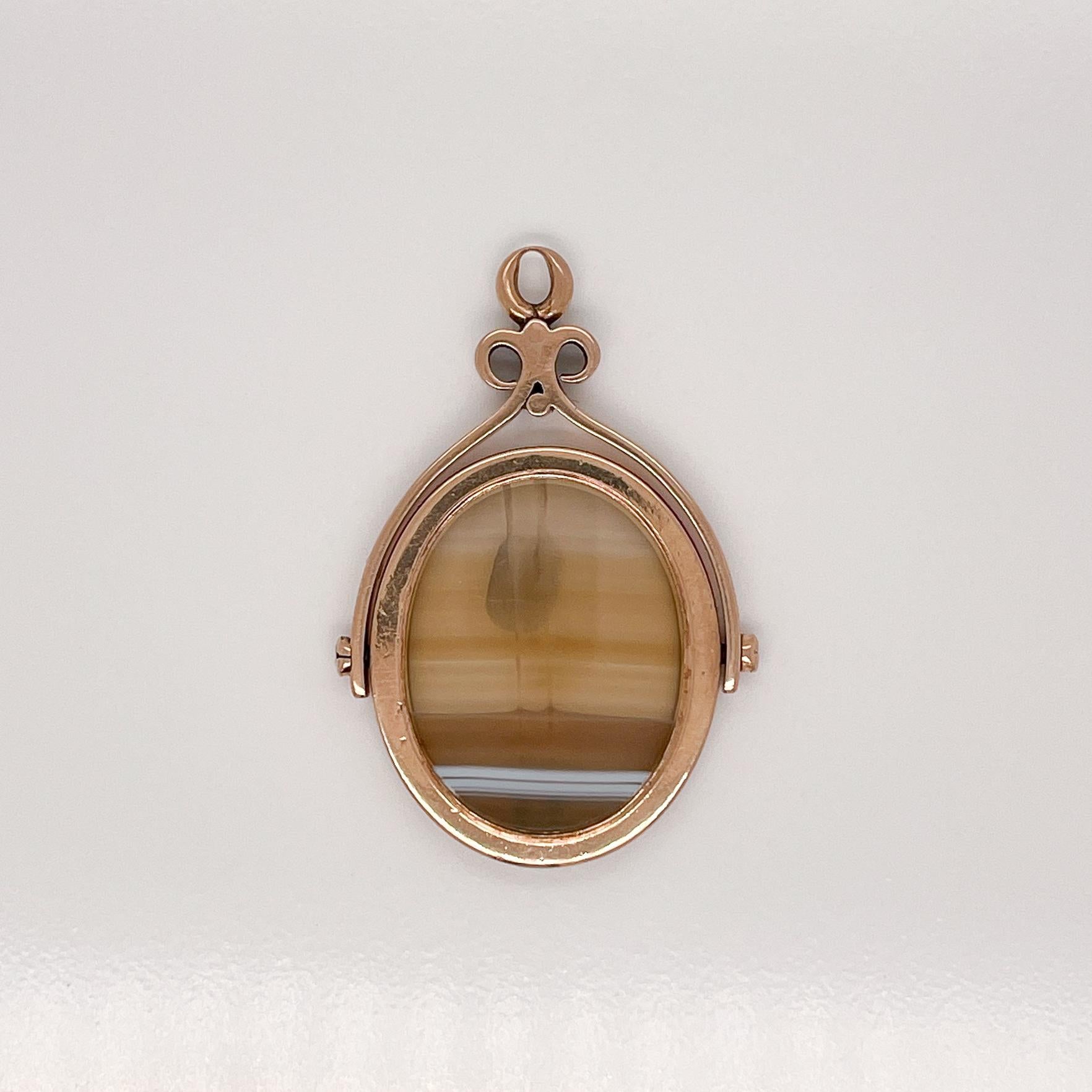 Un très beau pendentif à breloque en or antique et agate.

Avec une tranche ovale lisse d'agate sertie en or 14k et une charnière fixée de chaque côté pour que le charme puisse se retourner. 

Soutenu par un cadre en forme de volute avec une anse au