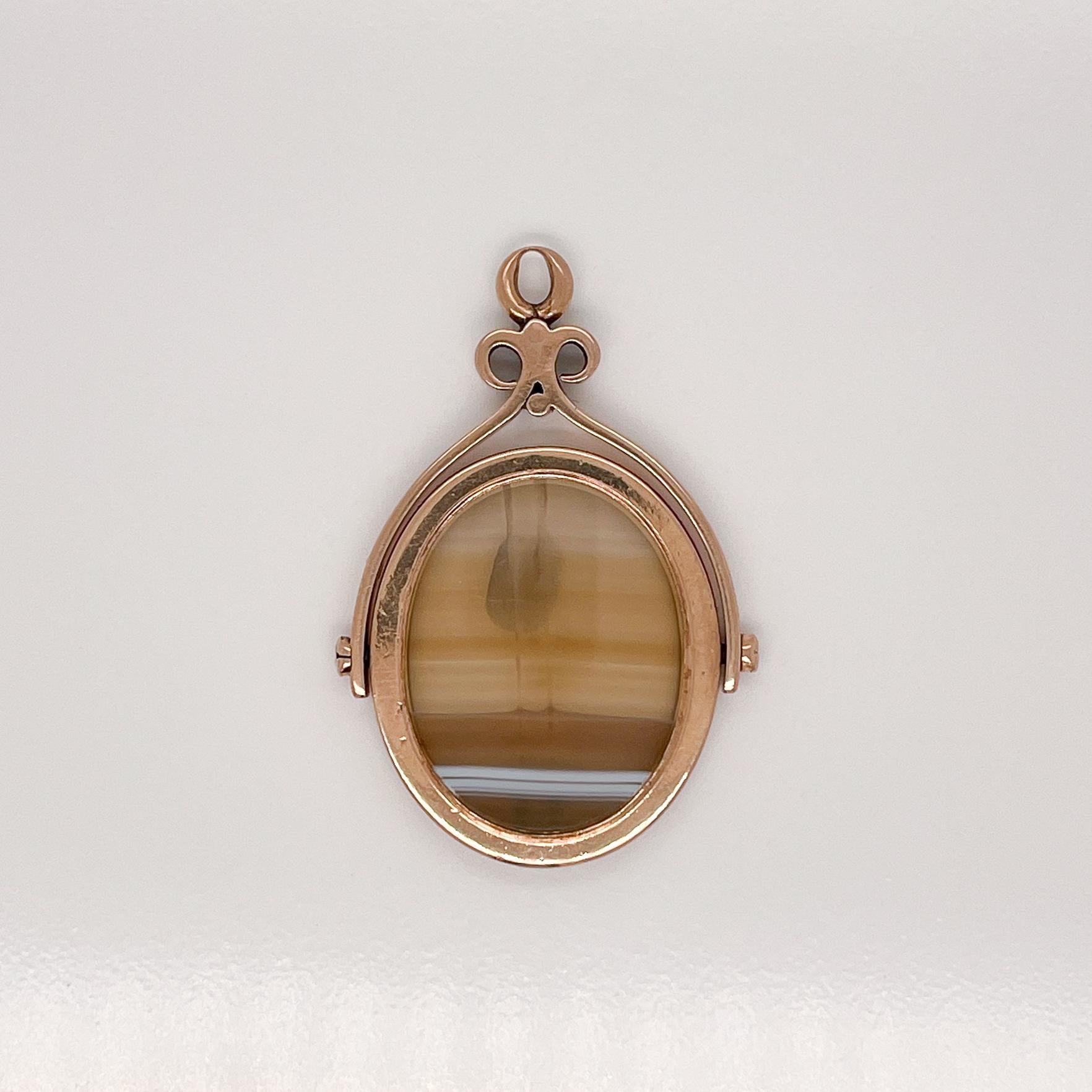 Un très beau pendentif à breloque en or antique et agate.

Avec une tranche ovale lisse d'agate sertie en or 14k et une charnière fixée de chaque côté pour que le charme puisse se retourner. 

Soutenu par un cadre en forme de volute avec une anse au