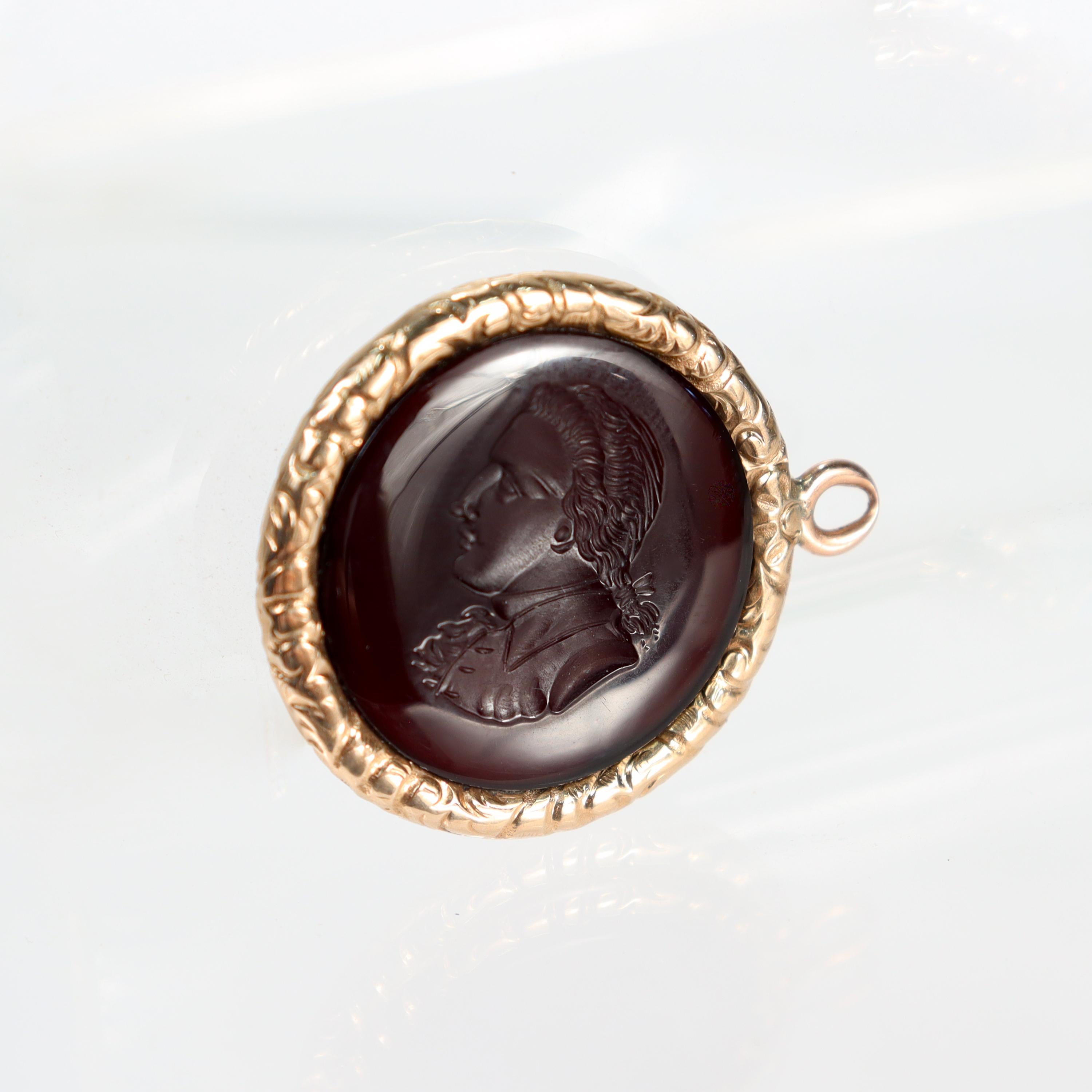 A fine intaglio pendant fob.

In 14k yellow gold. 

With an intaglio carved portrait of a man carved at the center a framed carnelian disc.

The patterned frame has a filigree pattern and is set with a bale that allows it to be worn on a necklace or