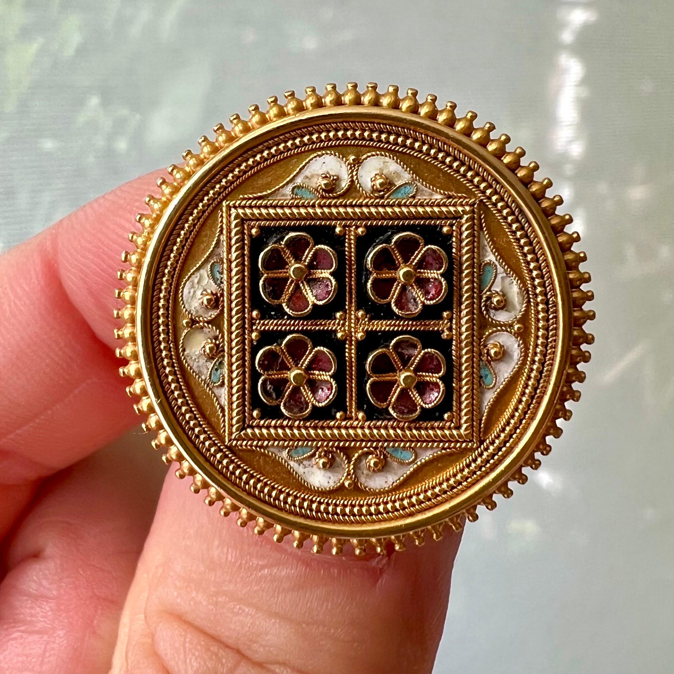 This is an antique round Etruscan Revival brooch made of 14 karat gold, decorated with an Art Nouveau floral motif of four flowers in a square with burgundy colored enamel petals. The brooch is inlaid with different colors of enamel, such as white,