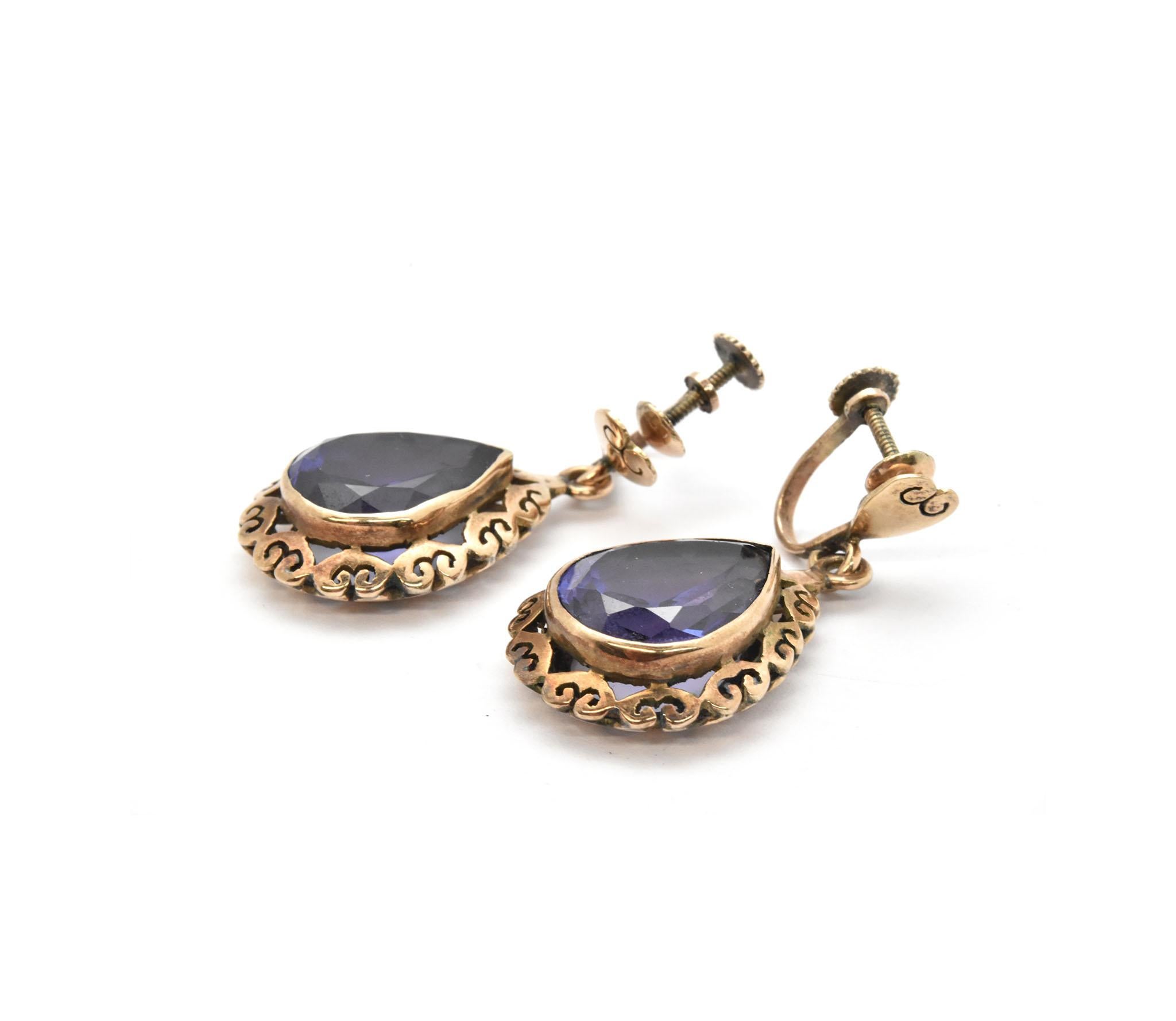 This antique pair of dangle earrings is made in 14k yellow gold with screw-tight, non-pierced backings. The earrings have bezel set pear-shaped amethyst gemstones surrounded by a hand-pierced heart shaped yellow gold decorated frame. The amethyst