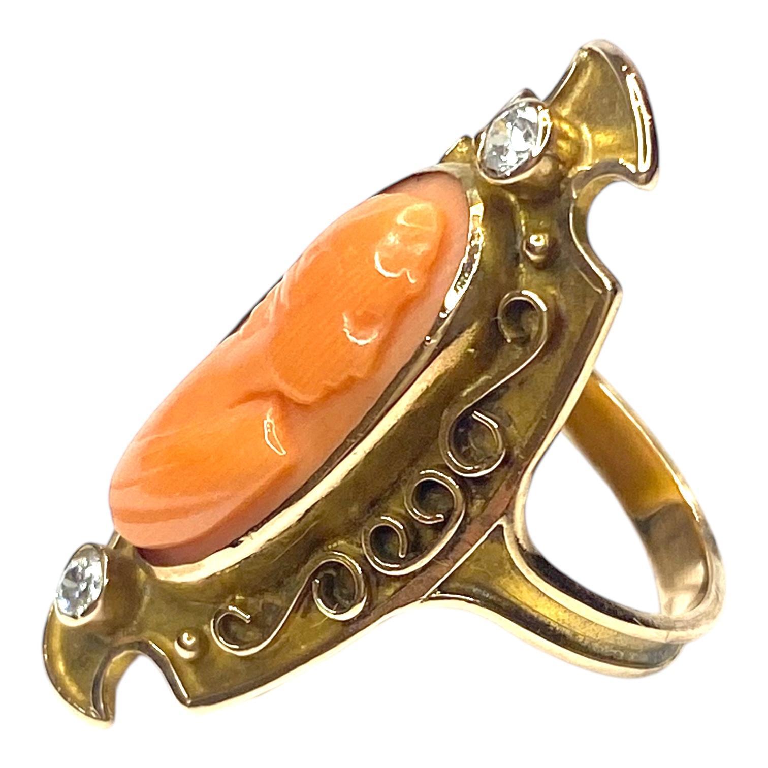 Crafted in 14K yellow gold, the ring features an oval coral cameo set in a gold bezel, accented by two bezel set diamonds in a low profile mounting with intricate scrollwork. 
2 Old Mine cut diamonds: 0.16ctw. Color: H-I, Clarity: