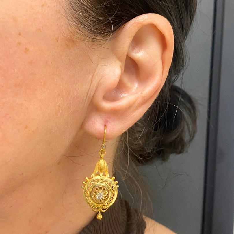 Paire de boucles d'oreilles en or antique, ornées de diamants taillés en rose.
Les pierres sont serties au centre d'un motif ajouré, entouré de détails en treillis d'or fin.
De petites perles dorées rehaussent le bord extérieur. Au-dessus de