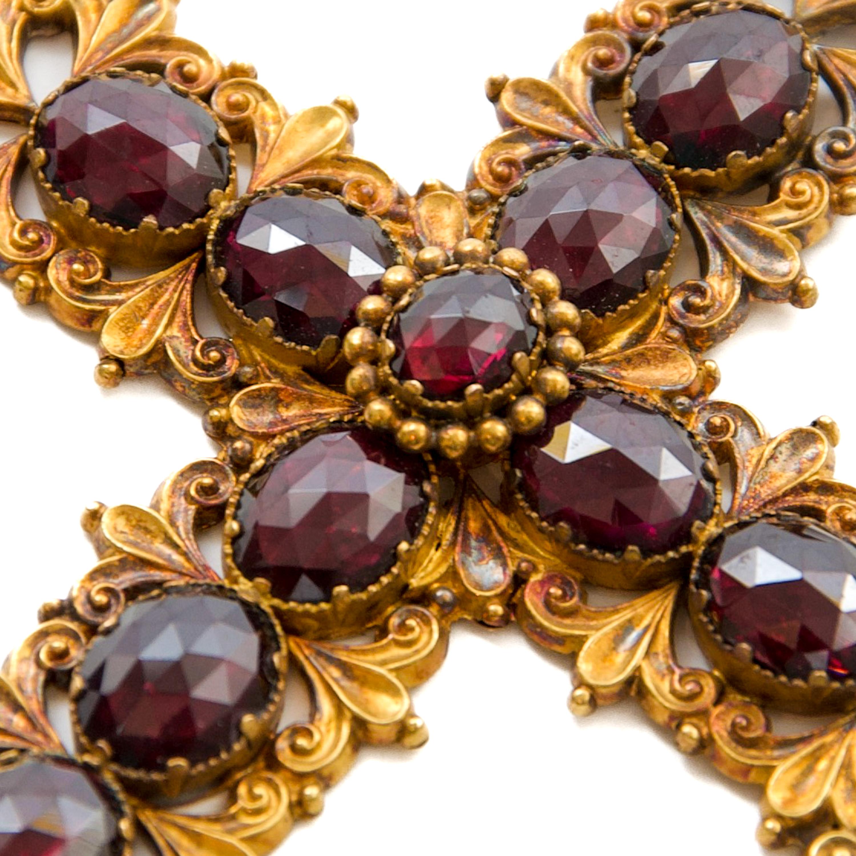 Questo splendido ciondolo a croce antico, realizzato in oro massiccio 14 carati, è un raffinato esempio di gioielleria ecclesiastica europea del XIX secolo con granati di Boemia riccamente sfaccettati in una cornice ornata di ispirazione barocca o