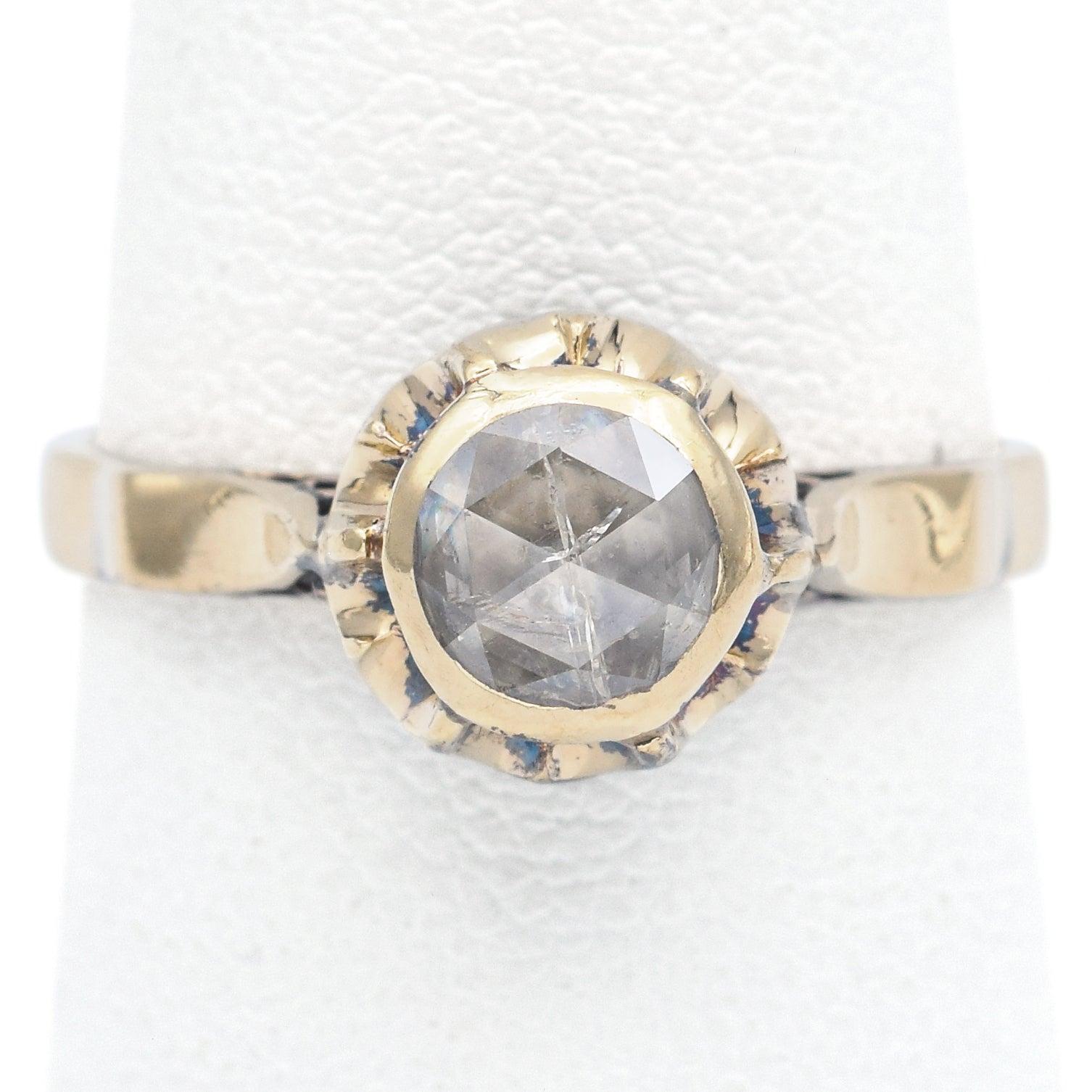 Un romántico anillo de compromiso antiguo con un diamante talla rosa, apreciado por su sutil brillo y su histórico facetado. El diamante está engastado en un bisel festoneado que se asemeja a un motivo floral, añadiendo un delicado encanto del viejo