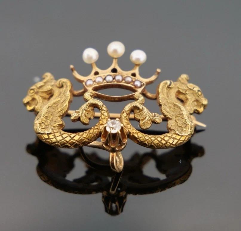 Diese auffällige antike Brosche ist aus massivem 18-karätigem Gelbgold gefertigt und zeigt zwei detailreiche Drachen, die eine königliche Krone flankieren, die mit natürlichen Saatperlen und einem Diamanten in Kesselfassung geschmückt ist. Eine