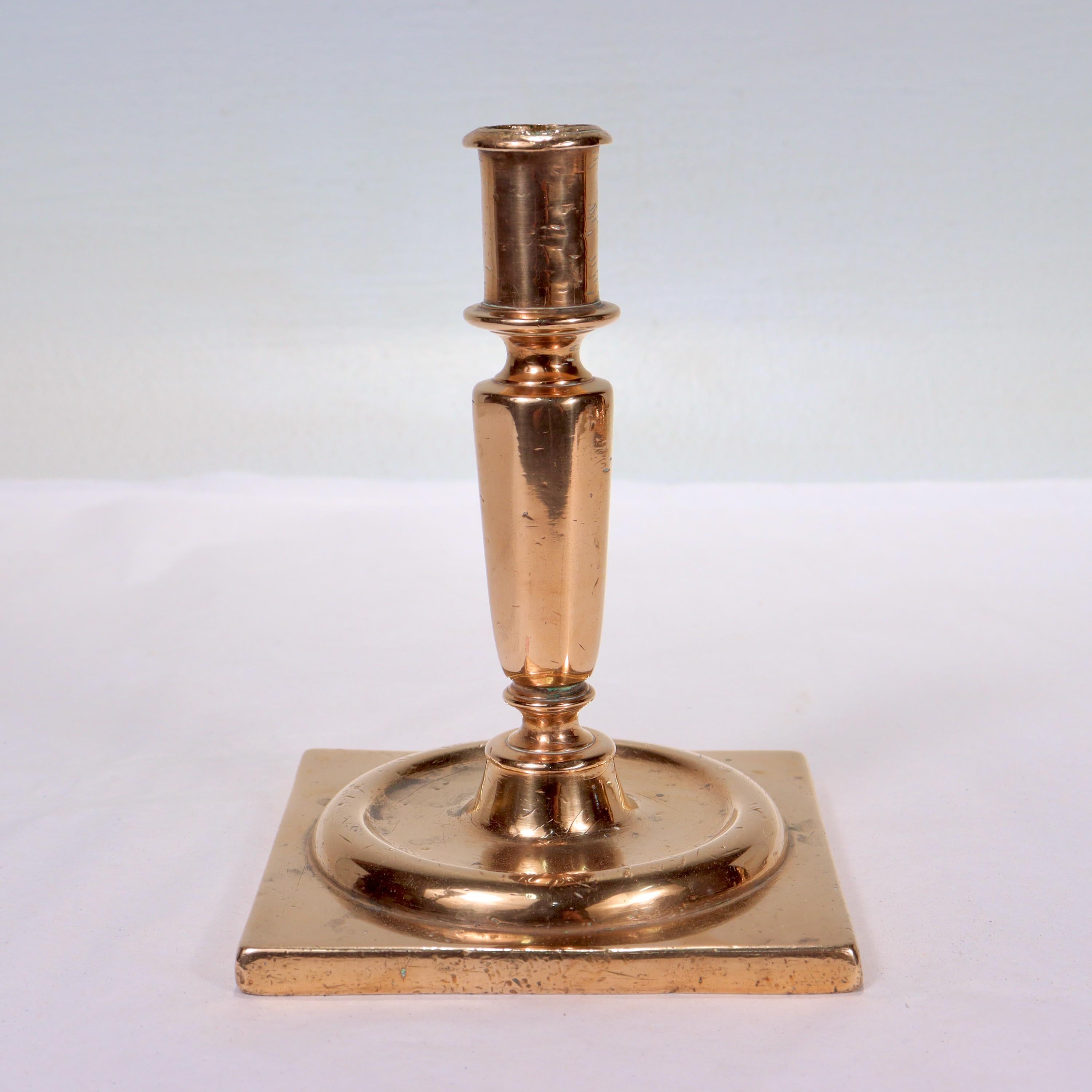 A fine antique bronze candlestick. 

Dating from the end of the 17th century.

Likely Dutch or English.

With a slightly orange hue and a wide square base supporting a tapered, faceted stem & cylindrical candle cup with a wax hole. The