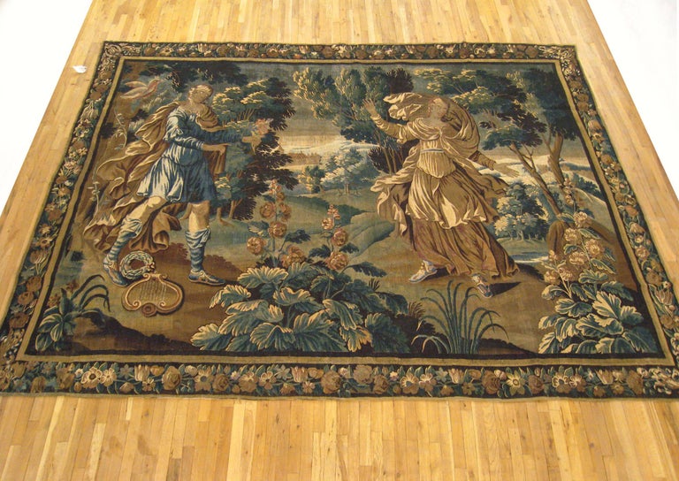 An antique 17th century Flemish mythological tapestry, size 9'1 H x 12'8 W, envisioning The Courtship of Apollo, with the Greek deity Apollo at left with his lyre and laurel garland, courting Daphne before her metamorphosis into a laurel tree. The