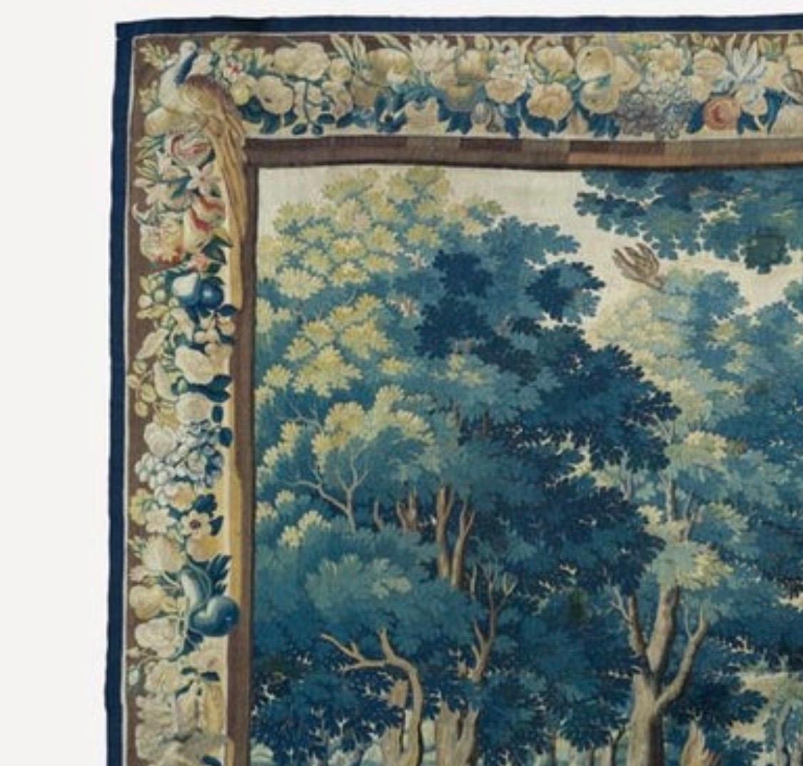 This is a gorgeous antique 17th century Flemish verdure landscape tapestry depicting a beautiful and rich summer scene of a countryside with lush trees and vegetation, with two figures in the foreground. The border features several different