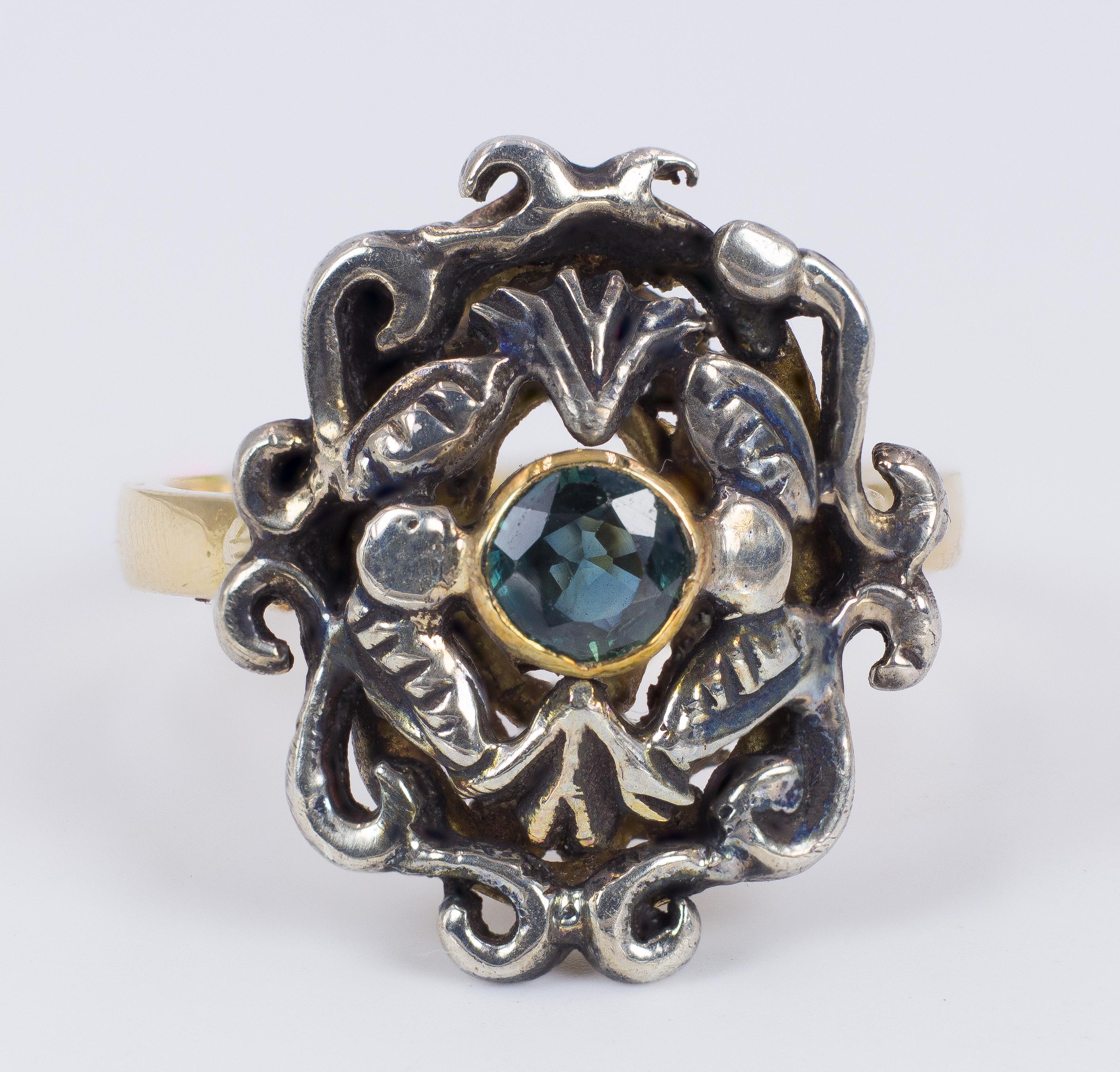 This antique ring, dating from the 1940s, features a very particular head, with an intricate wrought mount and with some spectacular foliate and flolar openworks. 
The central part of the ring is set with a blue topaz. 
The ring is modelled both in
