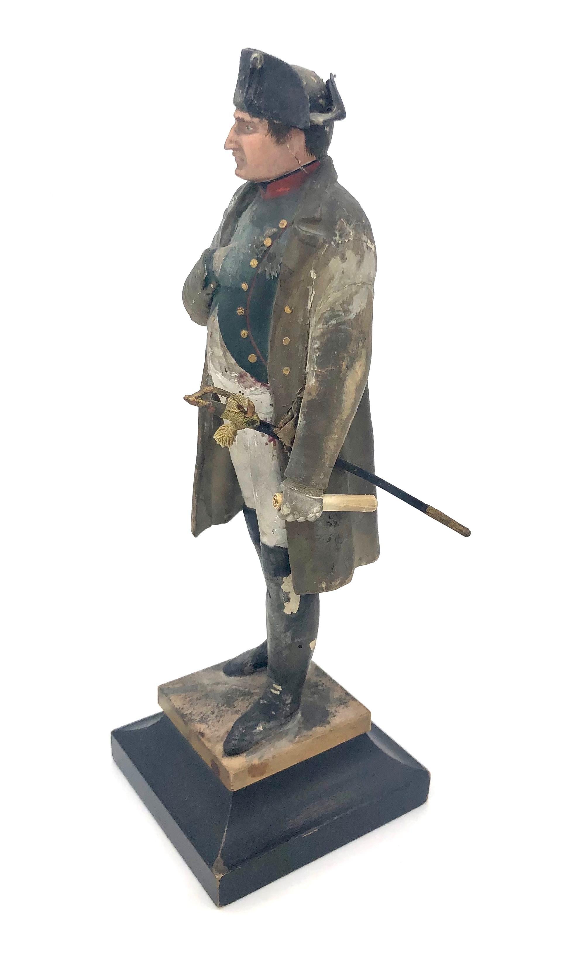 A statue of Napoleon with dagger in his favourite pose, his right hand in his unbuttoned waistcoat, his left hand holding a rolled up document. 
There are some losses to the back of the tricorn. Minor paint losses.