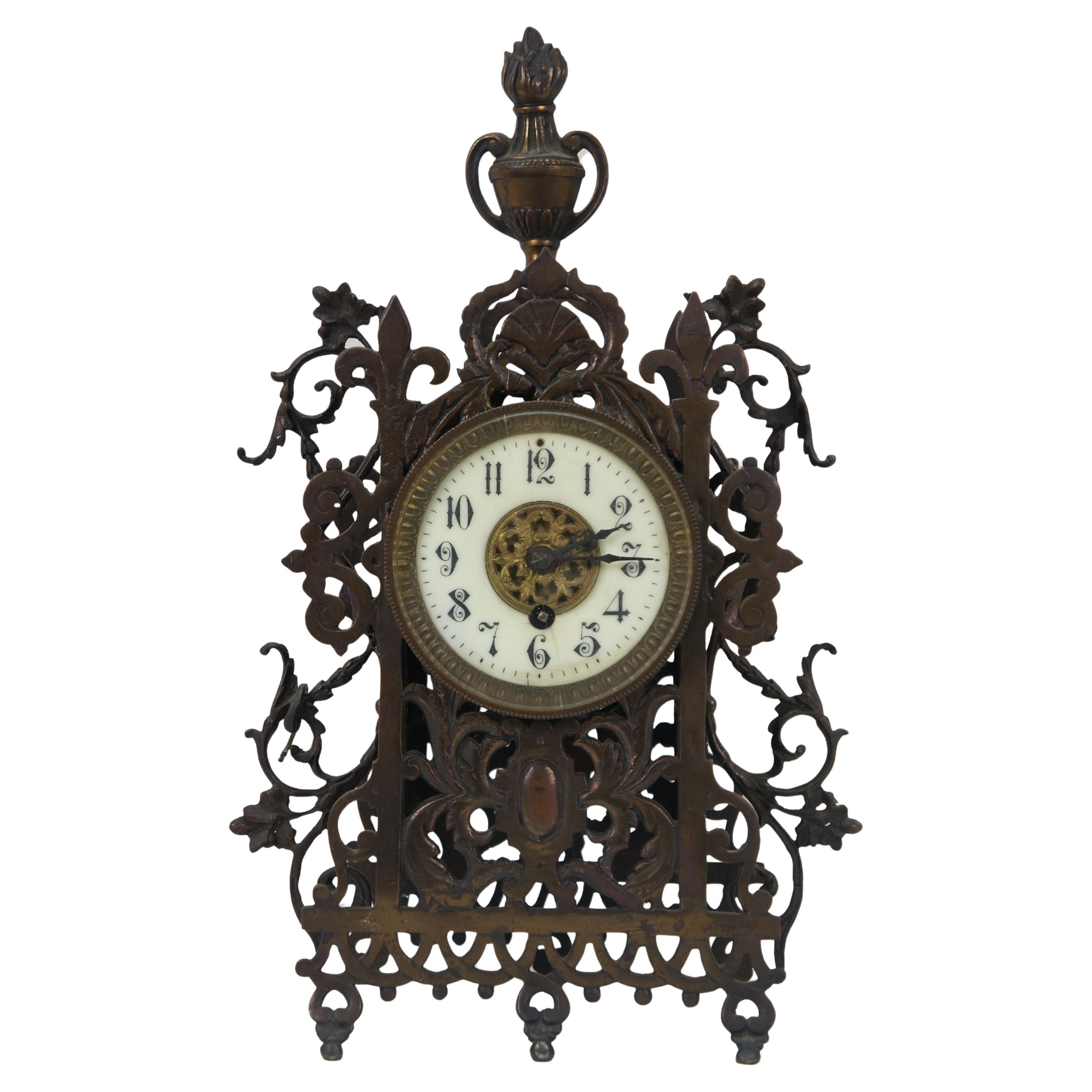 Antique Victorian c.1874 French bronze key wound mantel clock featuring an ornate scrolling pierced foliate design topped with fleur de lis and a flaming trophy urn. White face around a brass filigree medallion with black Arabic numerals and hands.