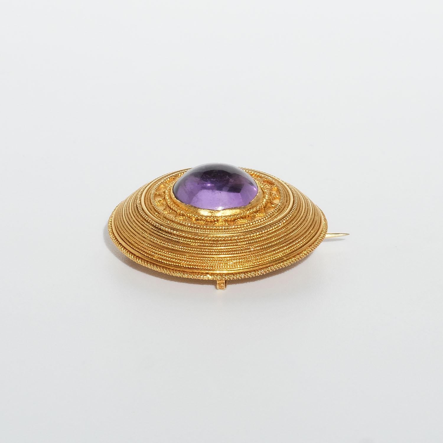 This antique 18 karat gold brooch is adorned with a center cabochon cut amethyst. The frame around the stone is beautifully patterned making it resemble a shield from ancient Greece.

The brooch is equipped with a loop which is why it can also be