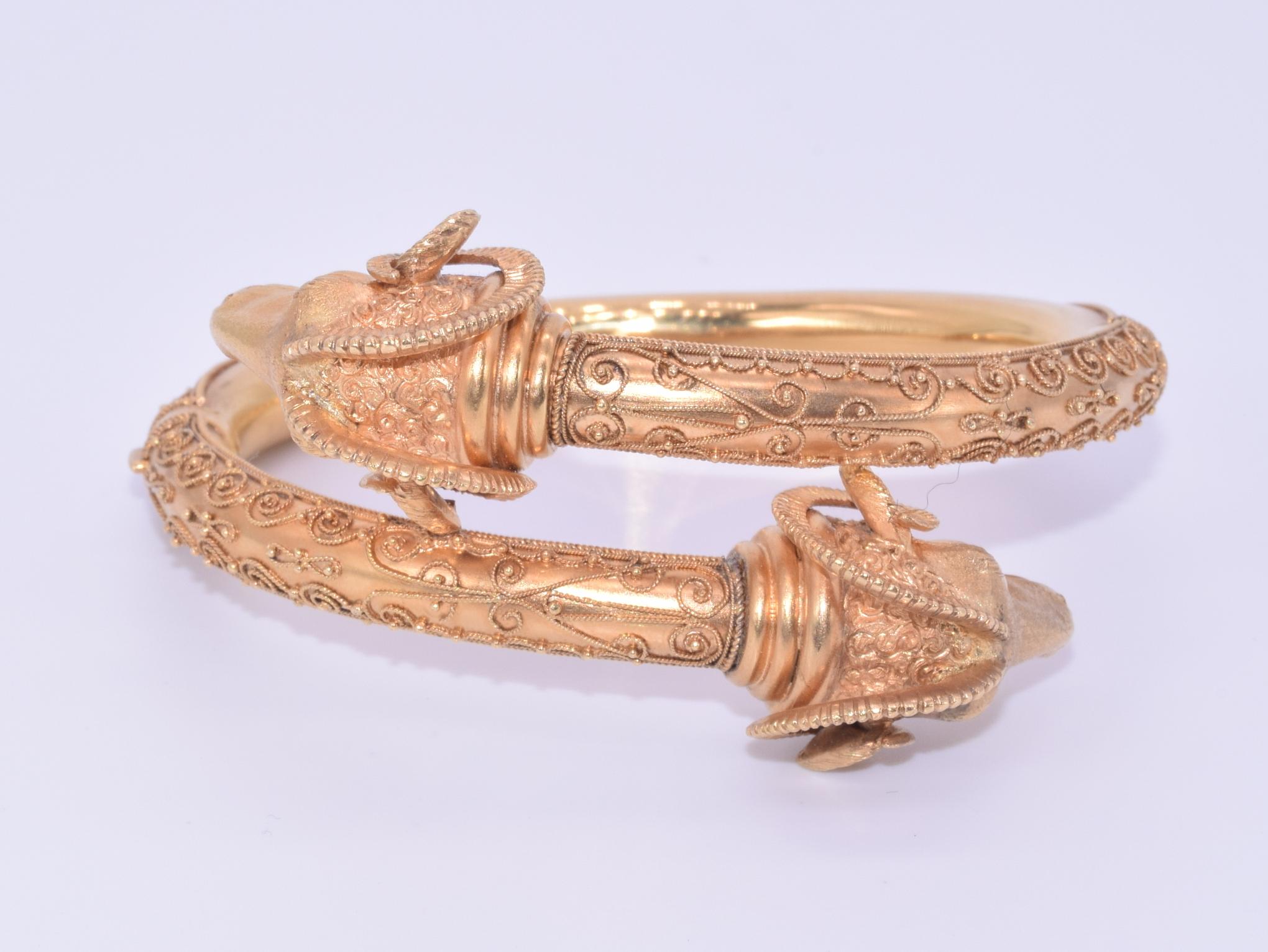 A hinged bypass bangle formed in 18 karat yellow gold features a pair of highly detailed ram's heads with granulation and scrolling wirework accents. Inner circumference approximately 6.5 inches. Each ram's head measures approximately 1 inch wide.
