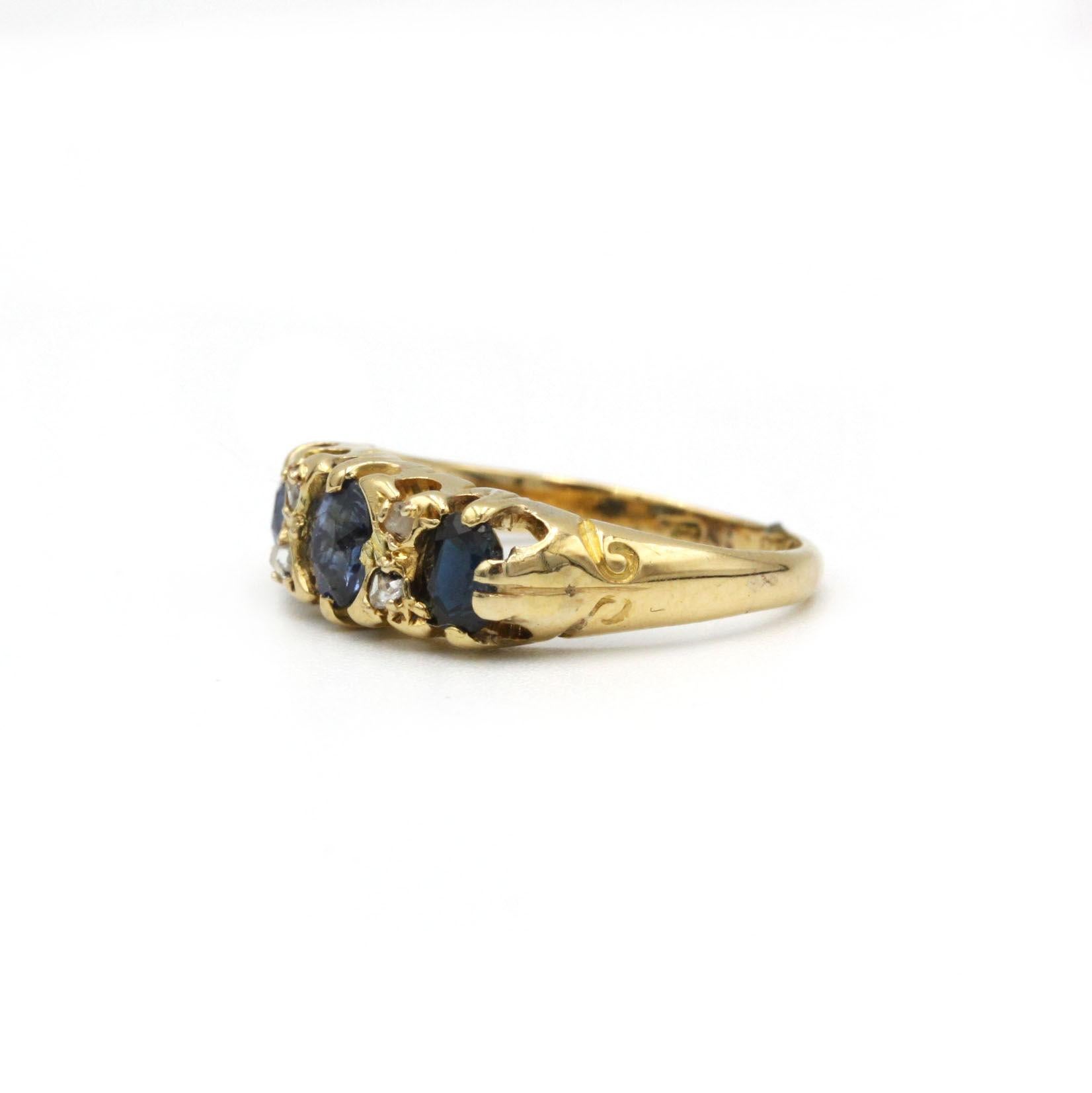 Cette exquise bague victorienne met en valeur trois saphirs bleus vibrants d'une valeur totale d'environ 1,00 carat, flanqués de quatre délicats diamants taillés en rose. Réalisée en or jaune 18 carats, la galerie à l'enroulement délicat et les