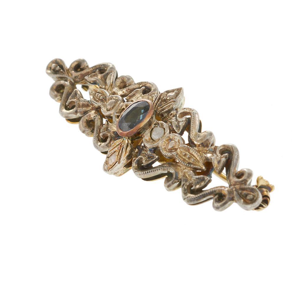 Un pezzo straordinario dell'epoca vittoriana, questa spilla antica è realizzata in oro giallo 18KT con una montatura in argento, con un intricato disegno di volute e motivi floreali. Al centro si trova uno zaffiro blu dal taglio ovale di .30 carati,