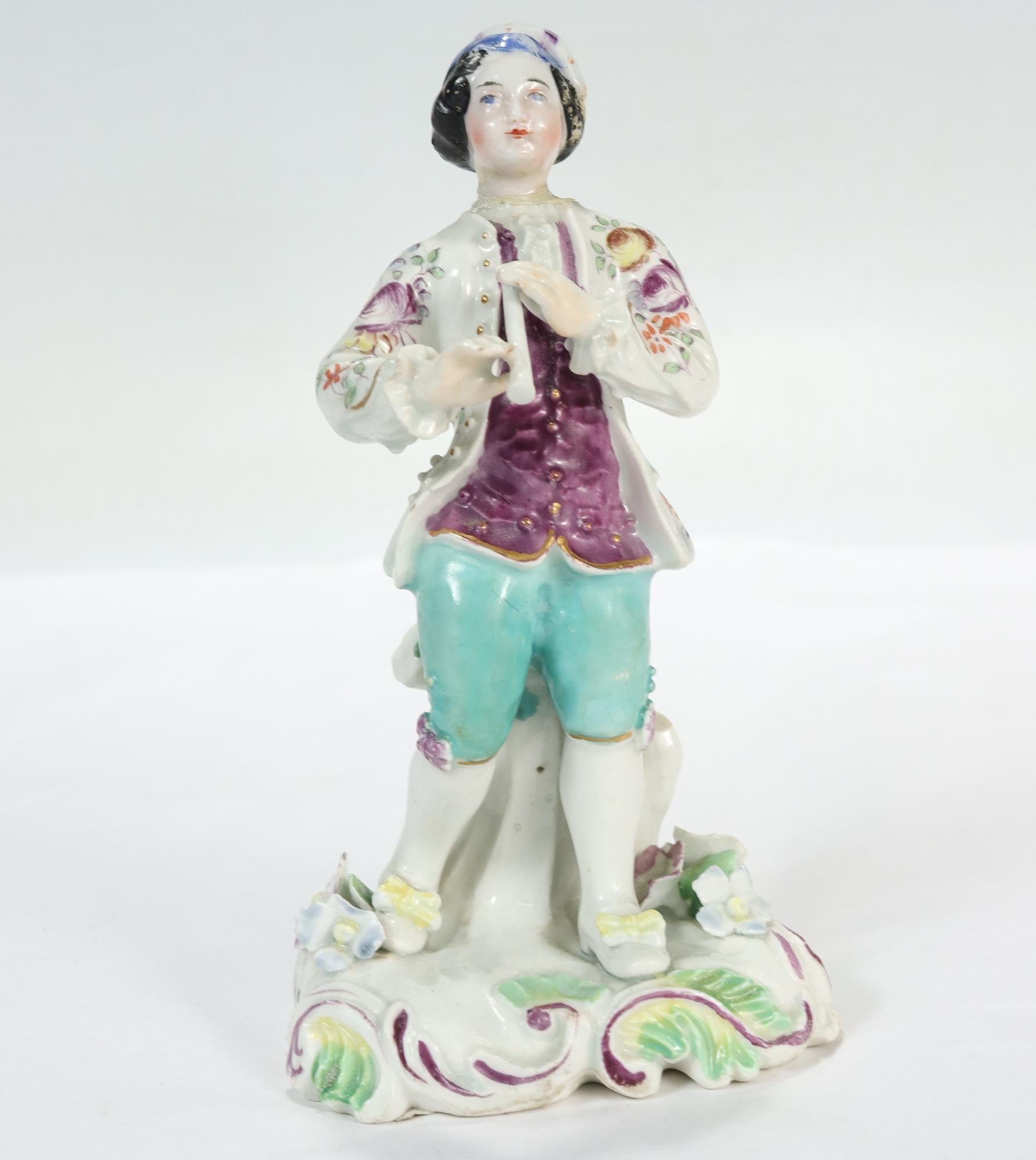 An antique English porcelain figurine.

By Bow. 

In the form of a boy clothed in 18th century garb and holding a flute.

We've noted losses to his flute, hands, hat, sleeves, and the flowers at his feet, as well as repairs to the neck and