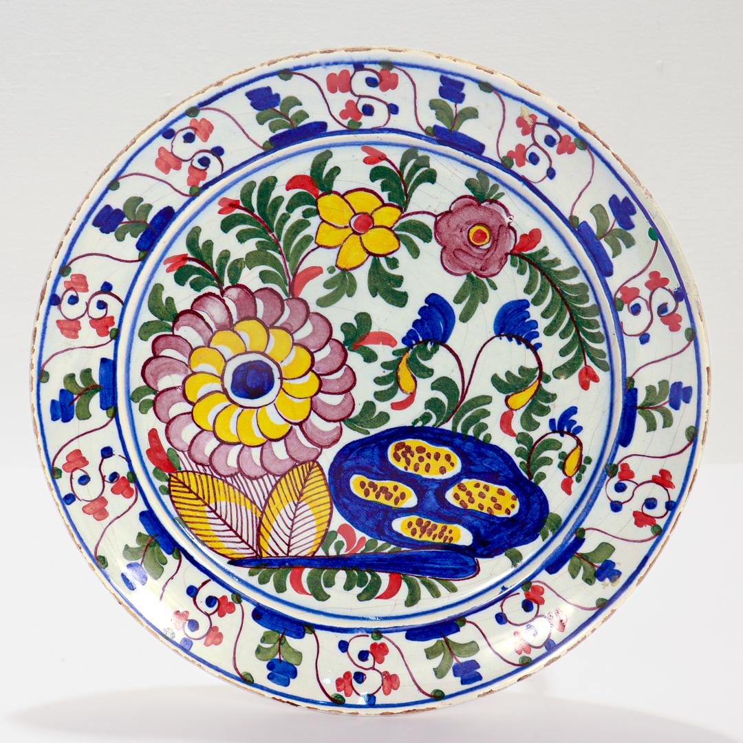 Une belle assiette ancienne en poterie hollandaise de Delft.

Avec un fond en porcelaine blanche richement décoré de fleurs et de feuillages bleus, verts, jaunes et orange.

La base porte la marque du fabricant De Klaauw, en brun sous glaçure.

Tout