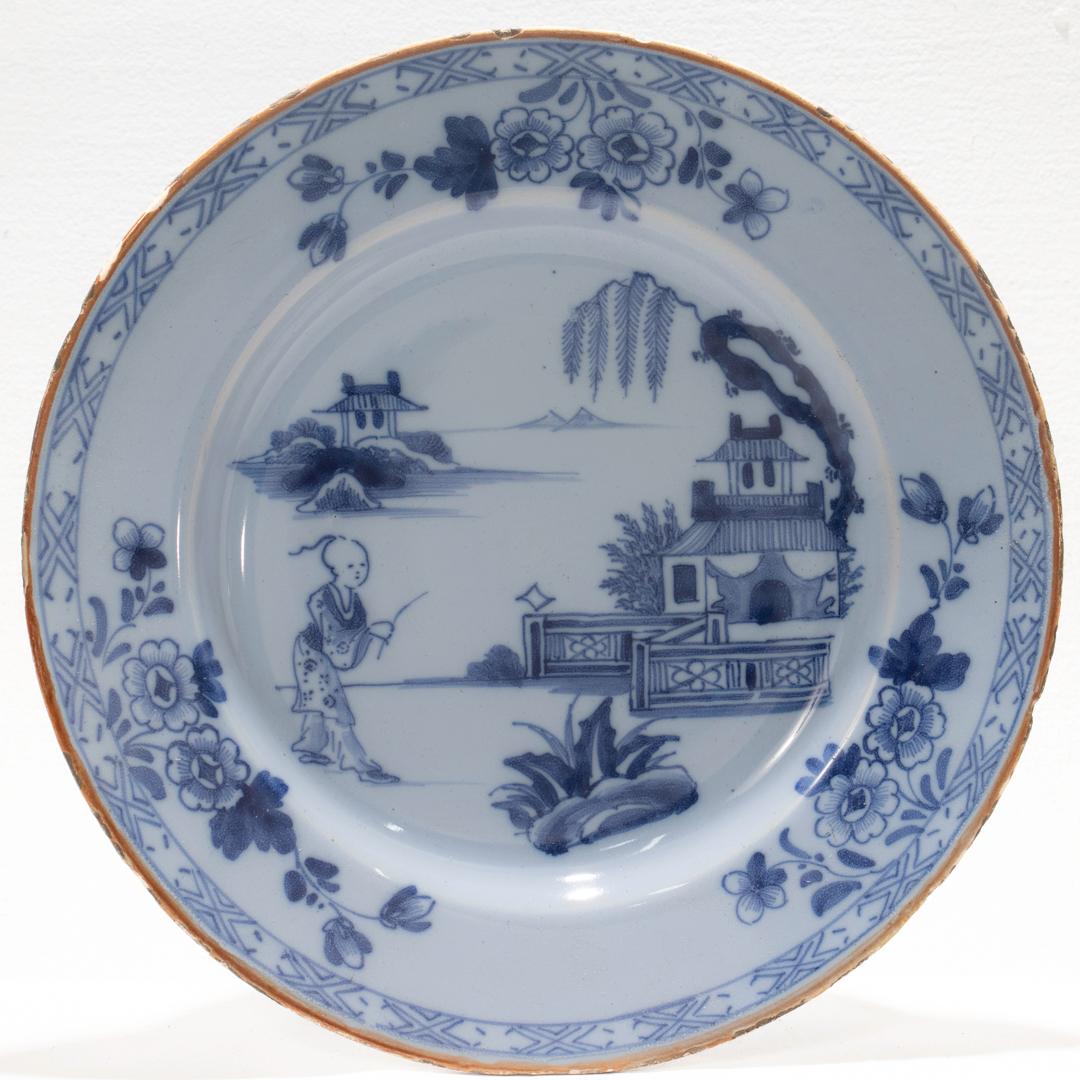 A fine antique Dutch Delft plate.

Depicting a man and fence in the foreground and a background with a house, a tree, an island with a building, and some distant mountains. 

The rim is decorated with floral accents and a geometric border.

Simply a