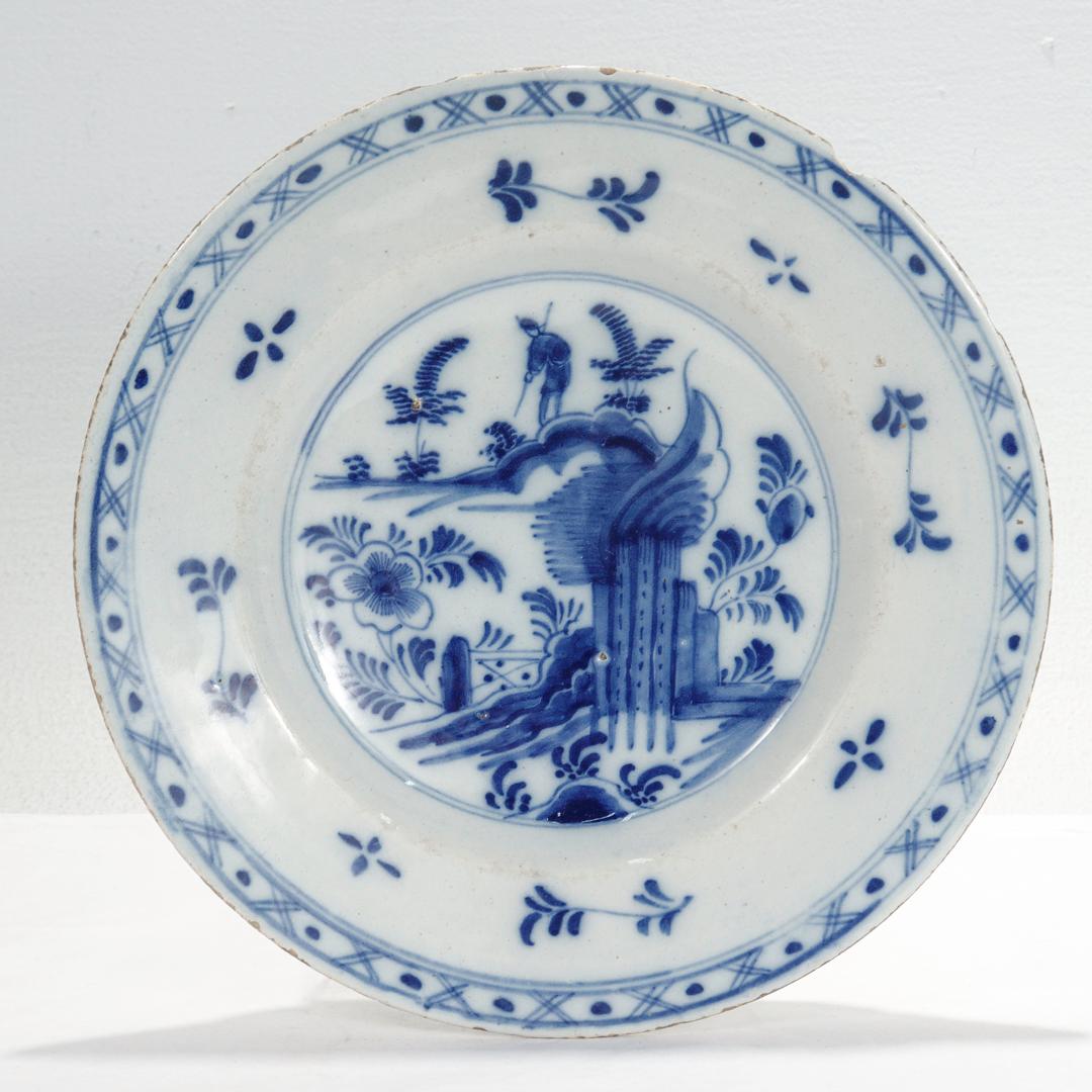 A fine antique 18th century Dutch Delft plate.

Depicting a garden in the foreground & a main in a landscape scene in the background. Surrounded by stylized floral sprigs and a dots & cross hatch decorated rim.

Simply a wonderful antique
