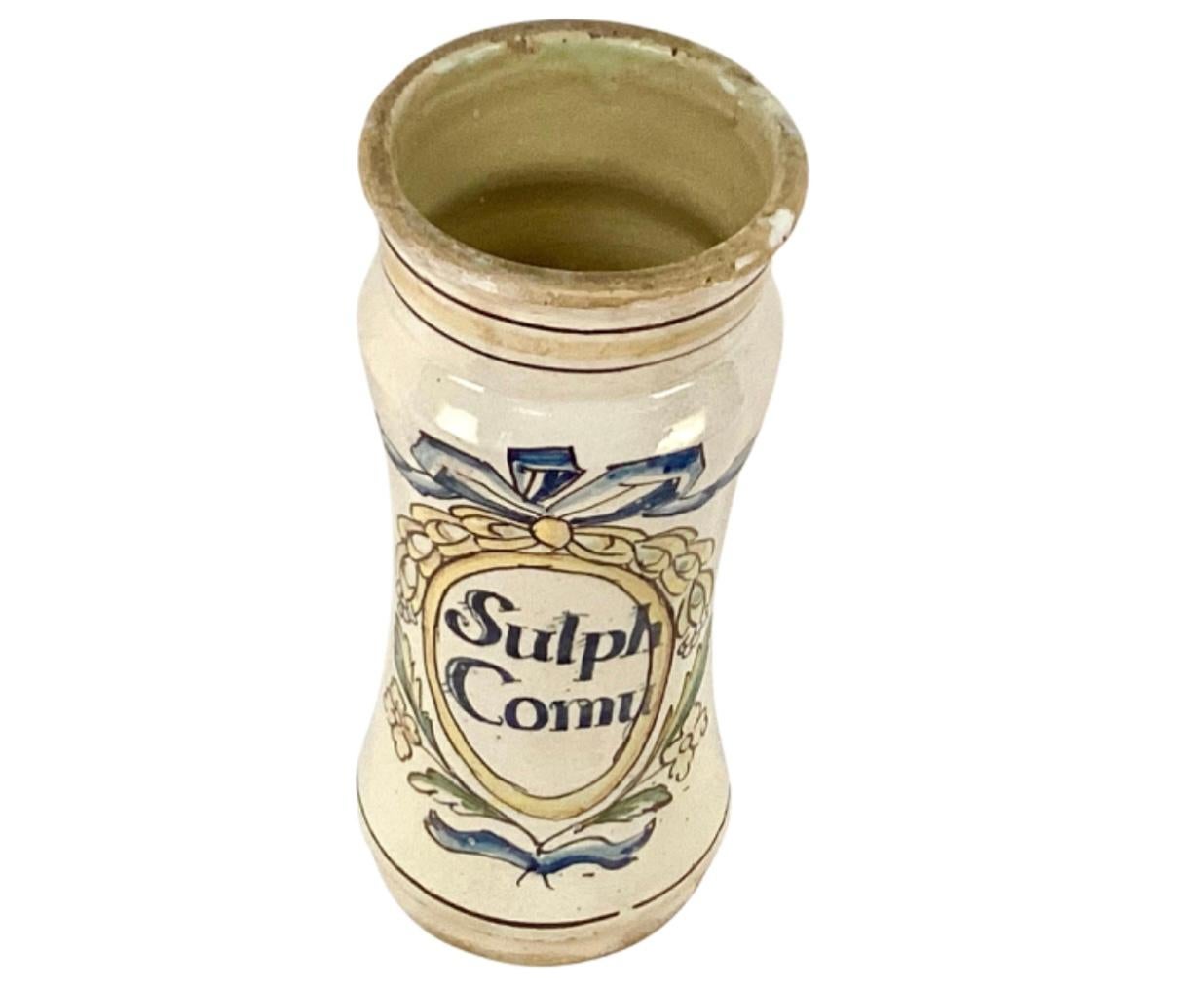 Tall vintage Italian ceramic apothecary jar. Has words 'Sulph Comu' on front. Floral and bow motif in colors of blue, yellow and green on off white background.
