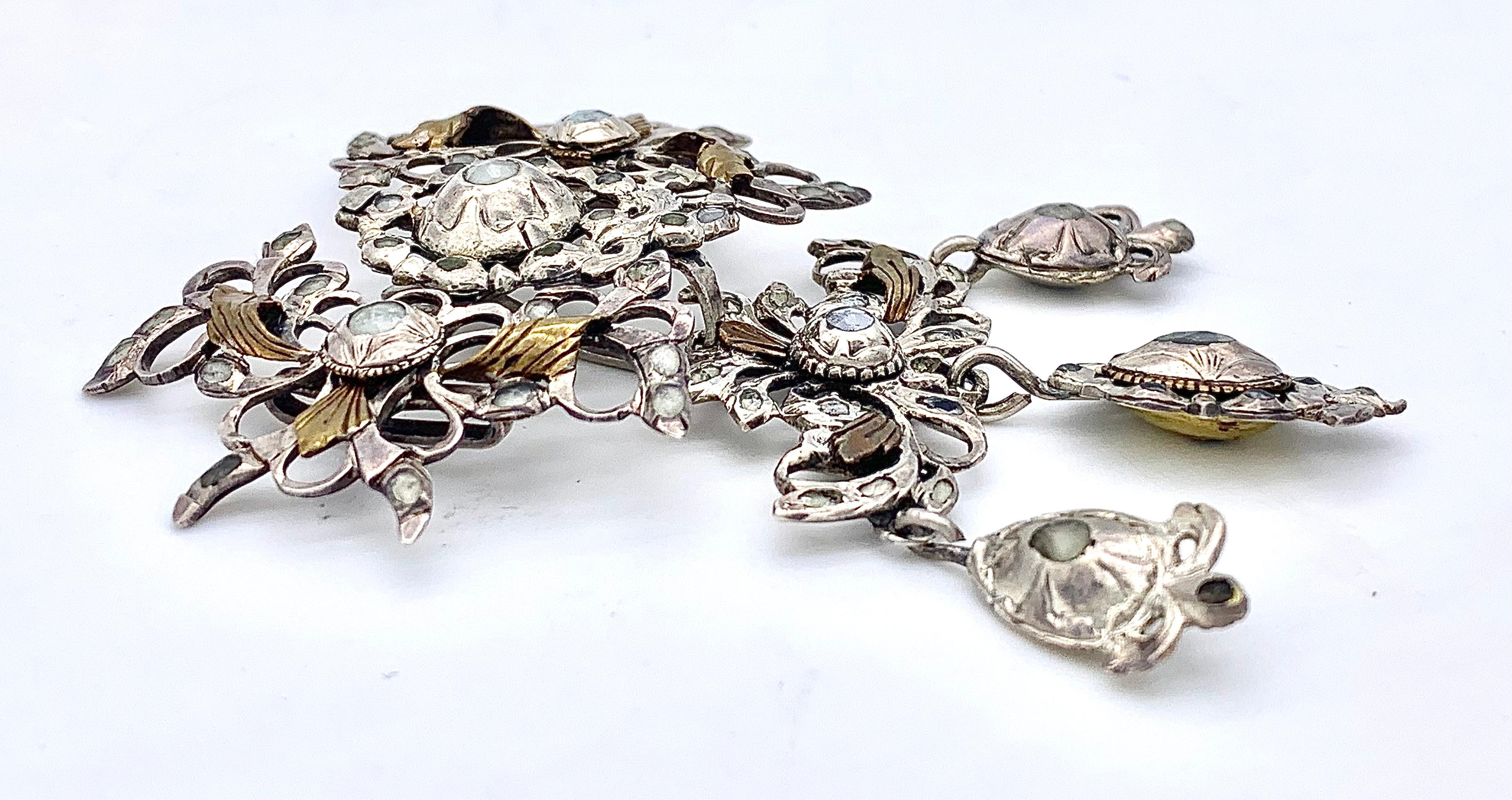Ce pendentif en argent de taille fine présente un motif percé avec des boutons de fleurs, des rubans, des nœuds et des feuilles dorées. Il est décoré de pierres en pâte dans des sertis fermés. Le plus grand serti fermé au centre de la lame présente