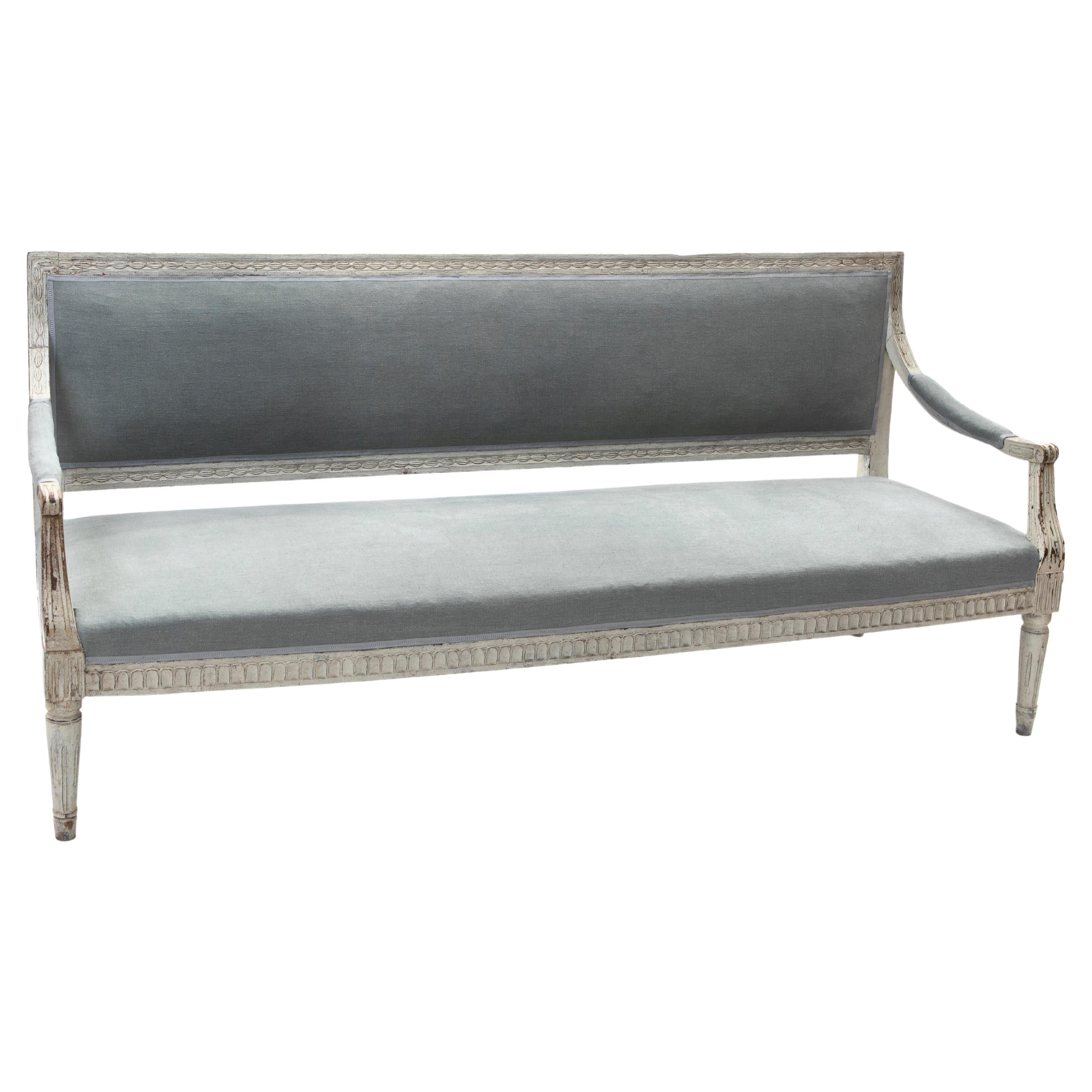 Swedish gustavian armed bench or sofa. 
Carved frame in light gray painted birchwood with natural age-related patina.

Features a long single seat with coil springs, upholstered with blue-gray sustainable hemp fabric from Les Créations de la