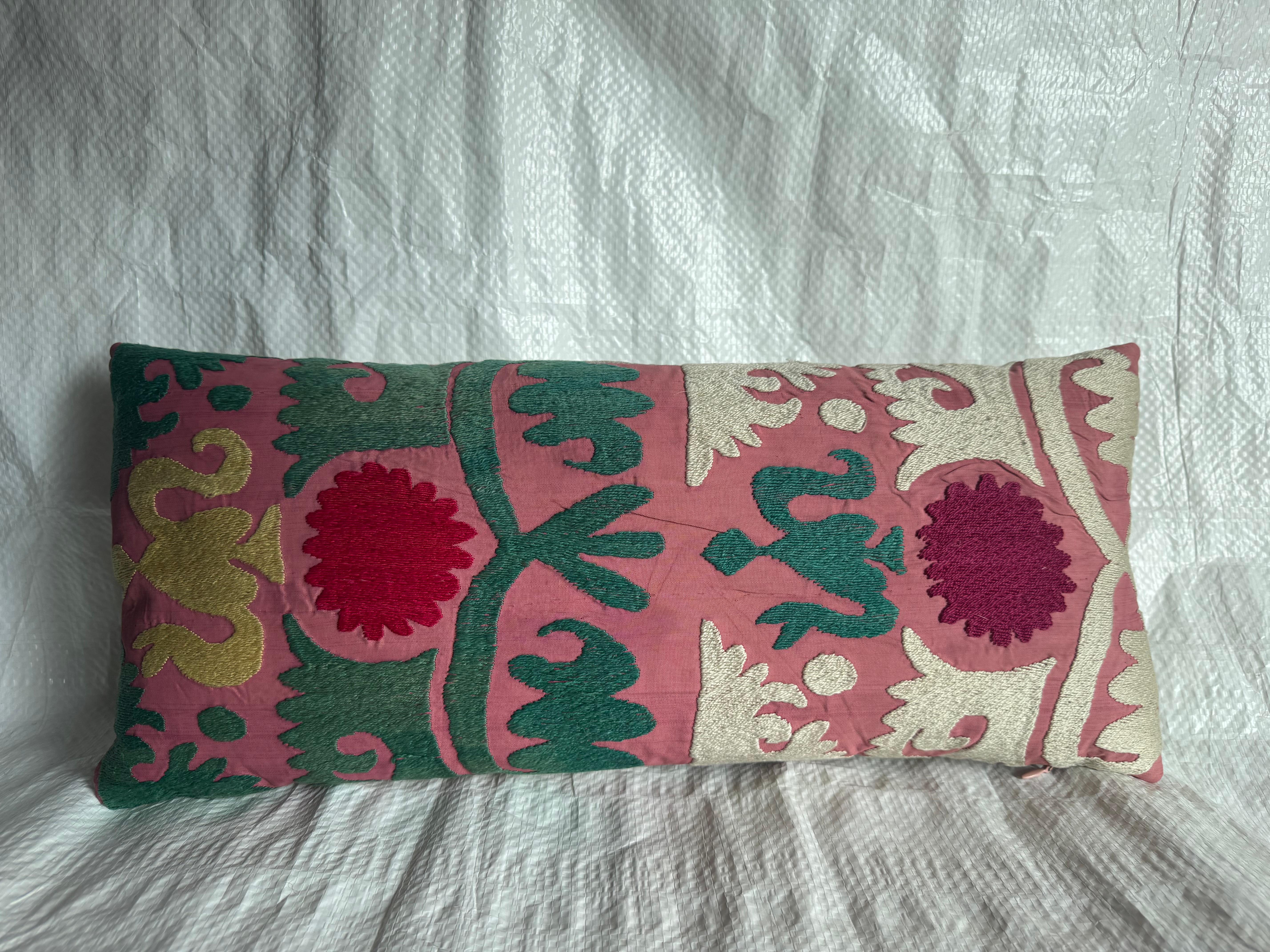 Uno squisito cuscino lombare Suzani antico, ricamato a mano in Asia centrale nei primi anni del 1900. Questo cuscino allungato presenta audaci medaglioni floreali e volute stilizzate, resi in vivaci tonalità di rosso, verde, avorio e oro su un fondo