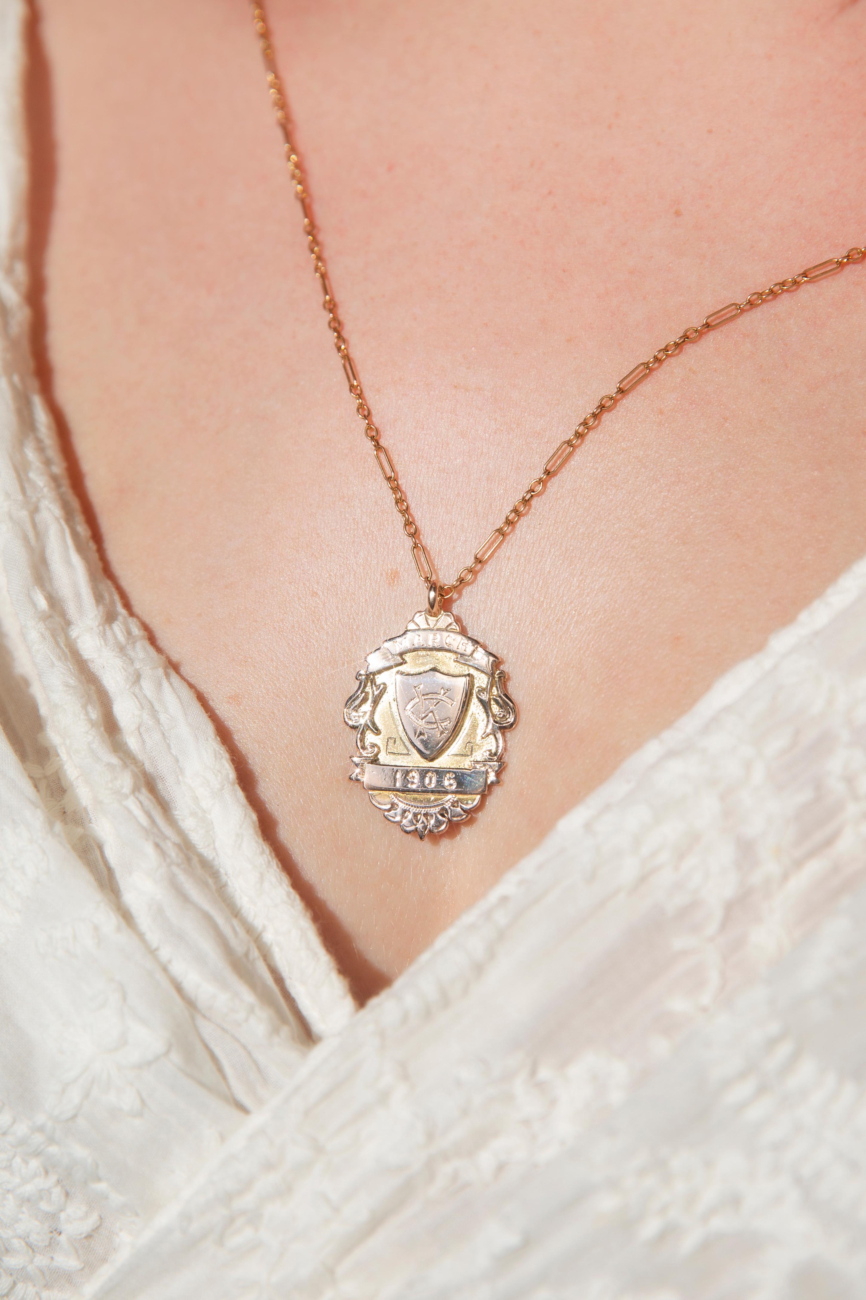 Crafted in 9 carat yellow gold, this lovely vintage antique pendant from 1906, during the Edwardian era, features a coat of arms with a shield at the centre inscribed with 