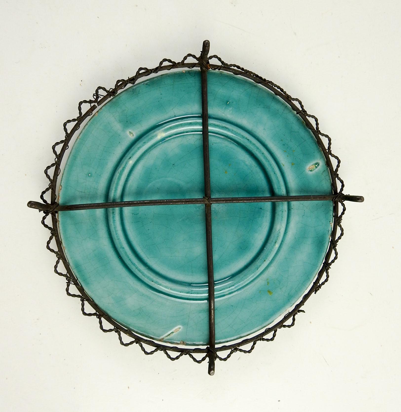 Turquoise majolica plate with boy catching a butterfly. Wrapped in wire with feet to use as a trivet or stand. No markings, small break in wire, surface wear to glaze.