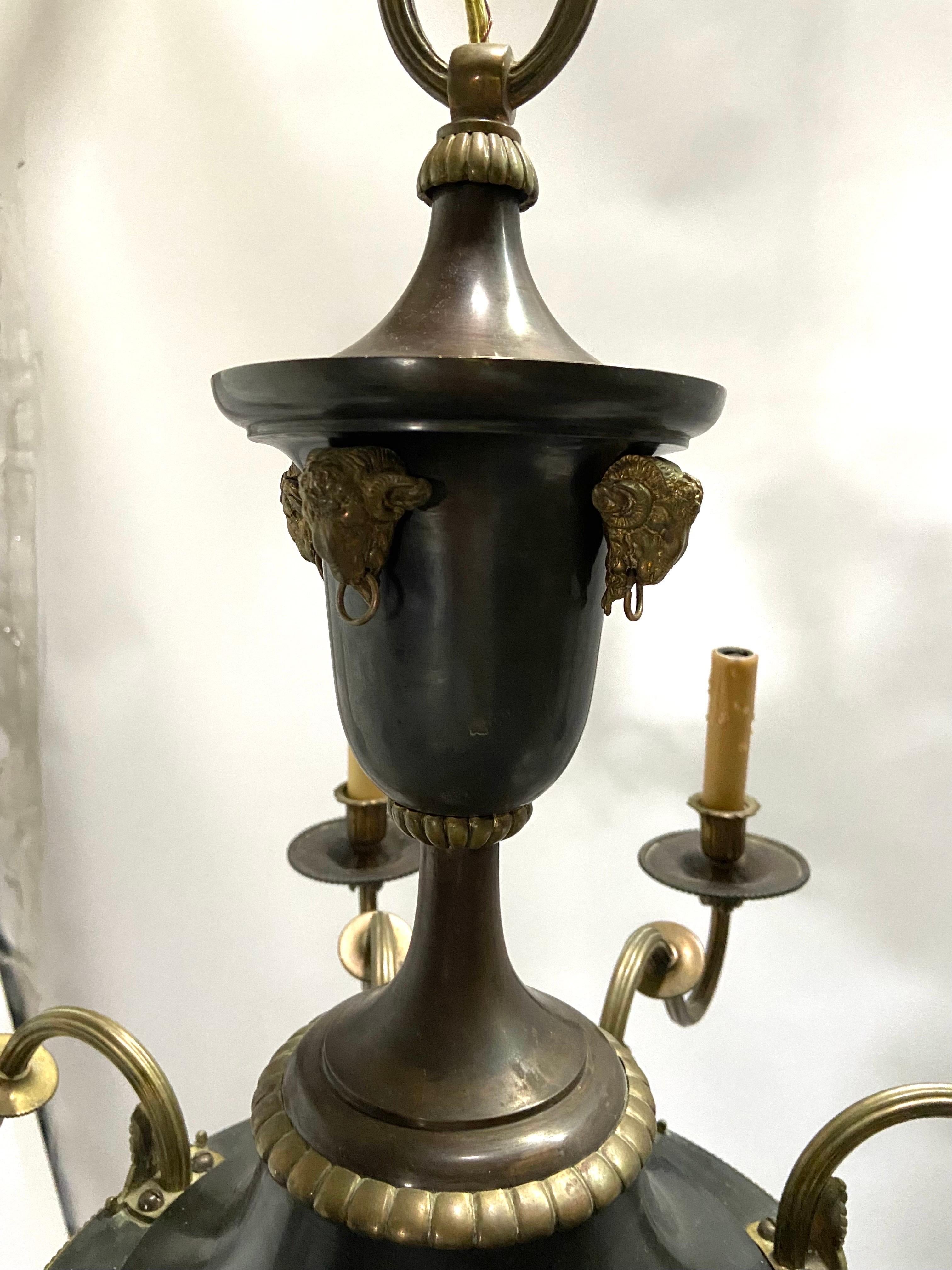 An antique, circa 1900 - 1920, French Empire style eight arm bronze chandelier with ram’s head details. This eight light chandelier has scroll and acanthus leaf details at each fluted and curved arm. The bobeches have a faceted underside and a
