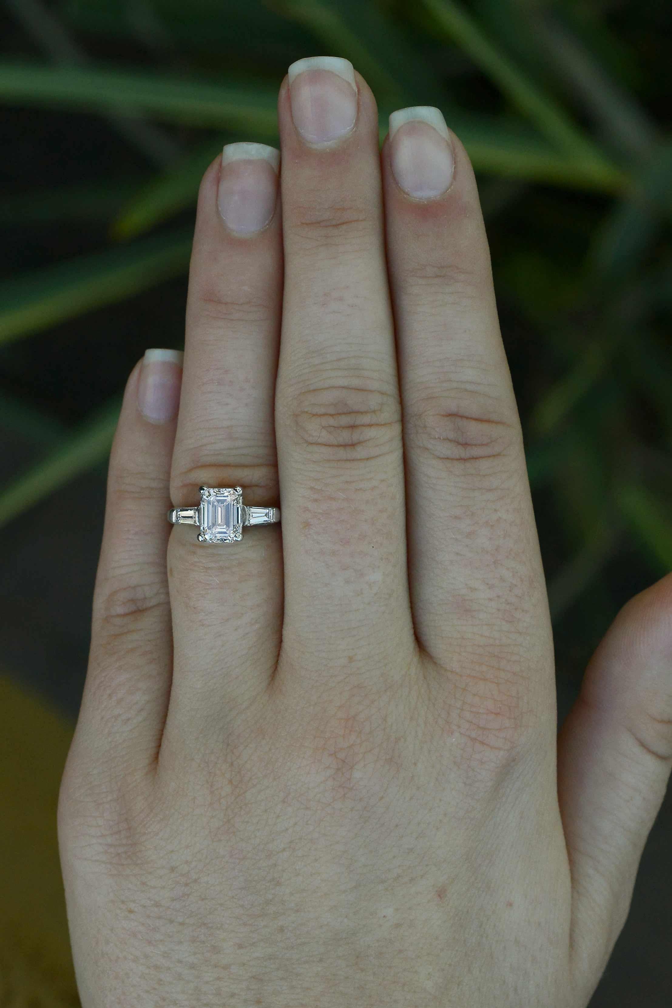 A subtle yet entrancing 3 stone Art Deco antique engagement ring, styled after Tiffany & Co. centered by a GIA certified 1.50 carat emerald cut diamond showing near flawless clarity and excellent symmetry. Graced with a scintillating near-colorless