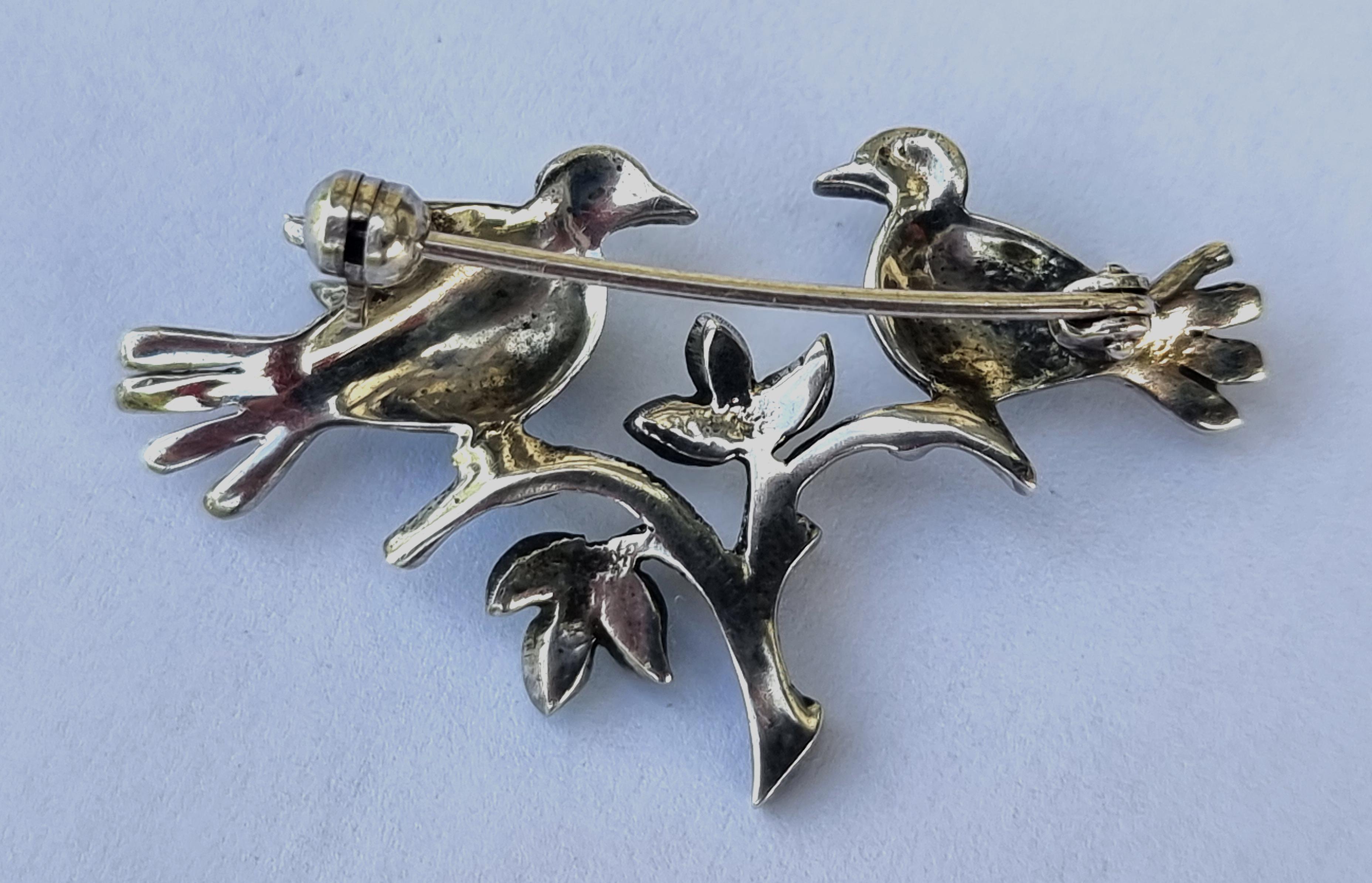 
Diese entzückende, wunderschön gearbeitete Silberbrosche aus den 1920er Jahren (nicht gestempelt, aber geprüft) zeigt ein Paar exotischer Liebesvögel auf einem belaubten Stiel. Es handelt sich um ein feines, delikates Exemplar, das von Hand mit