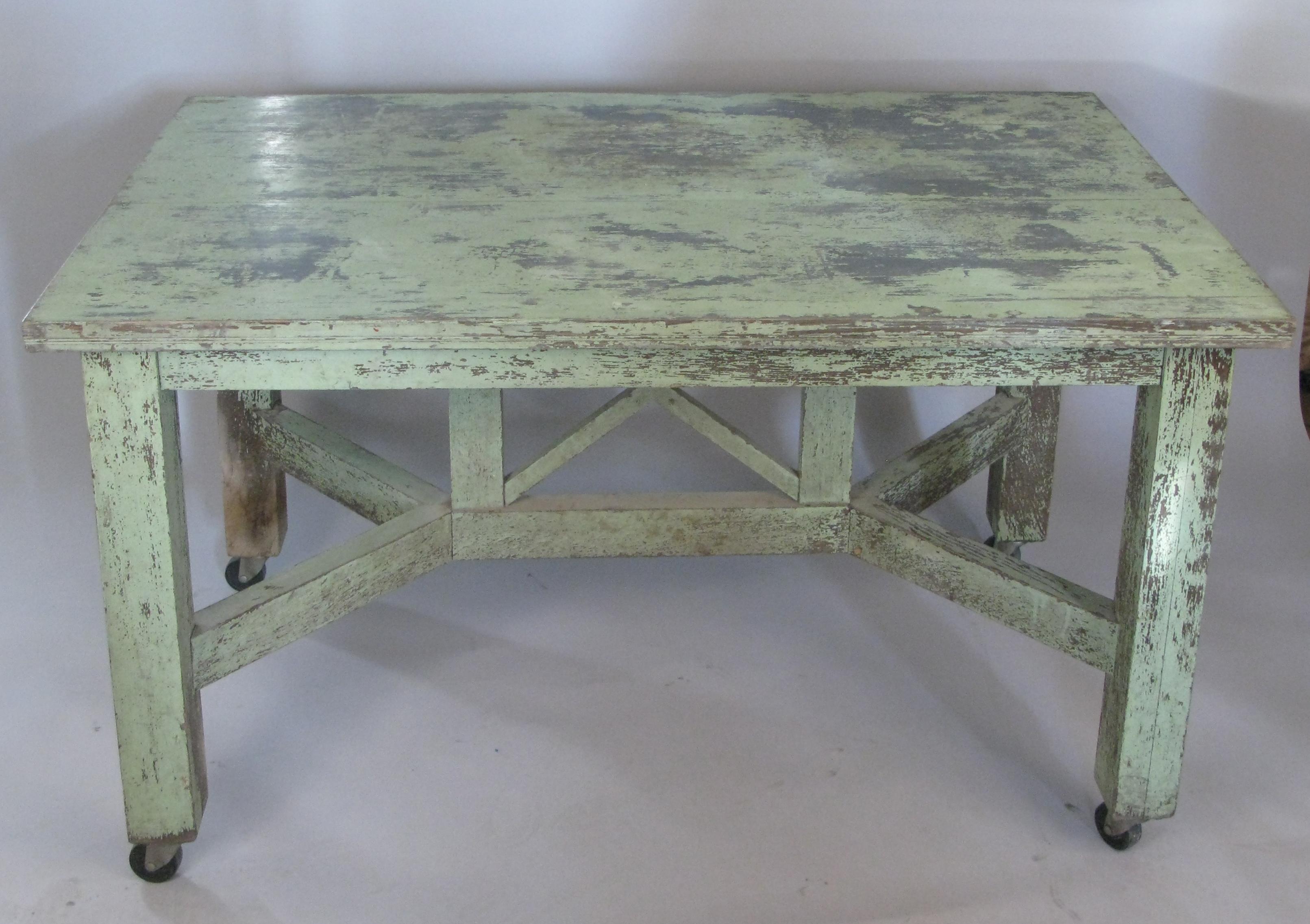 A very handsome antique 1940s country table with the remains of its original painted finish in green. the table has a very interesting base with an X-brace in the center, and is raised on casters, which could be removed. one side of the table has