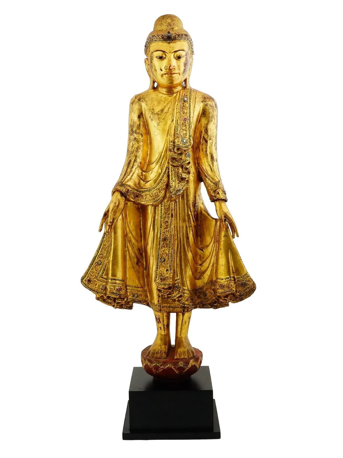 Statue antique de Bouddha debout dans la pose de l'appel de la pluie. De Birmanie, Mandalay, 19e siècle. La statue est en bois laqué doré et incrusté de détails en verre. Elle représente le Bouddha à l'expression sereine, debout sur un piédestal, et