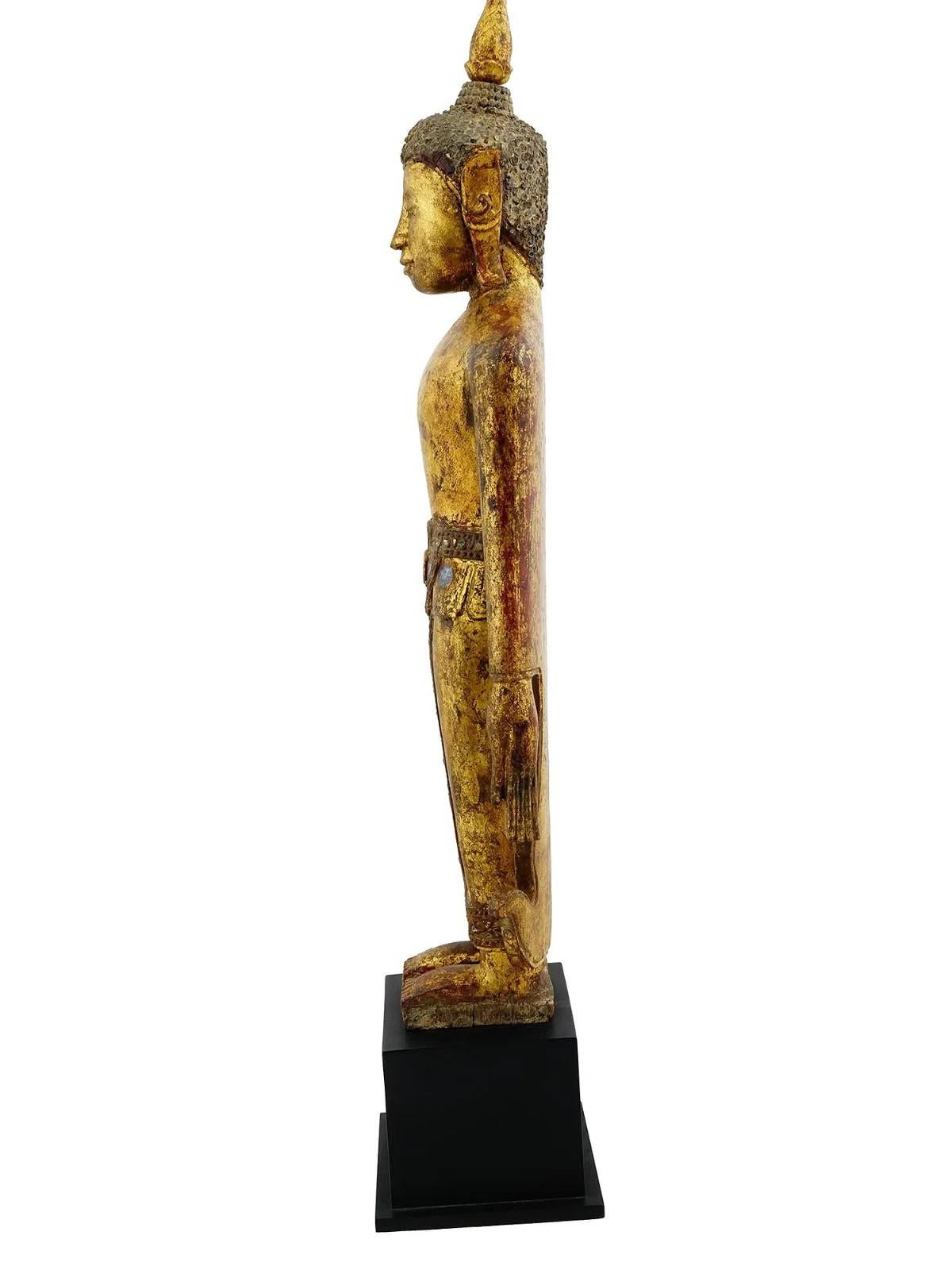 Statue antique de Bouddha debout dans la pose de l'appel de la pluie. De Birmanie, Mandalay, 19e siècle. La statue est réalisée en bois laqué doré et incrustée de détails en verre. Elle représente le Bouddha avec une expression sereine, debout sur