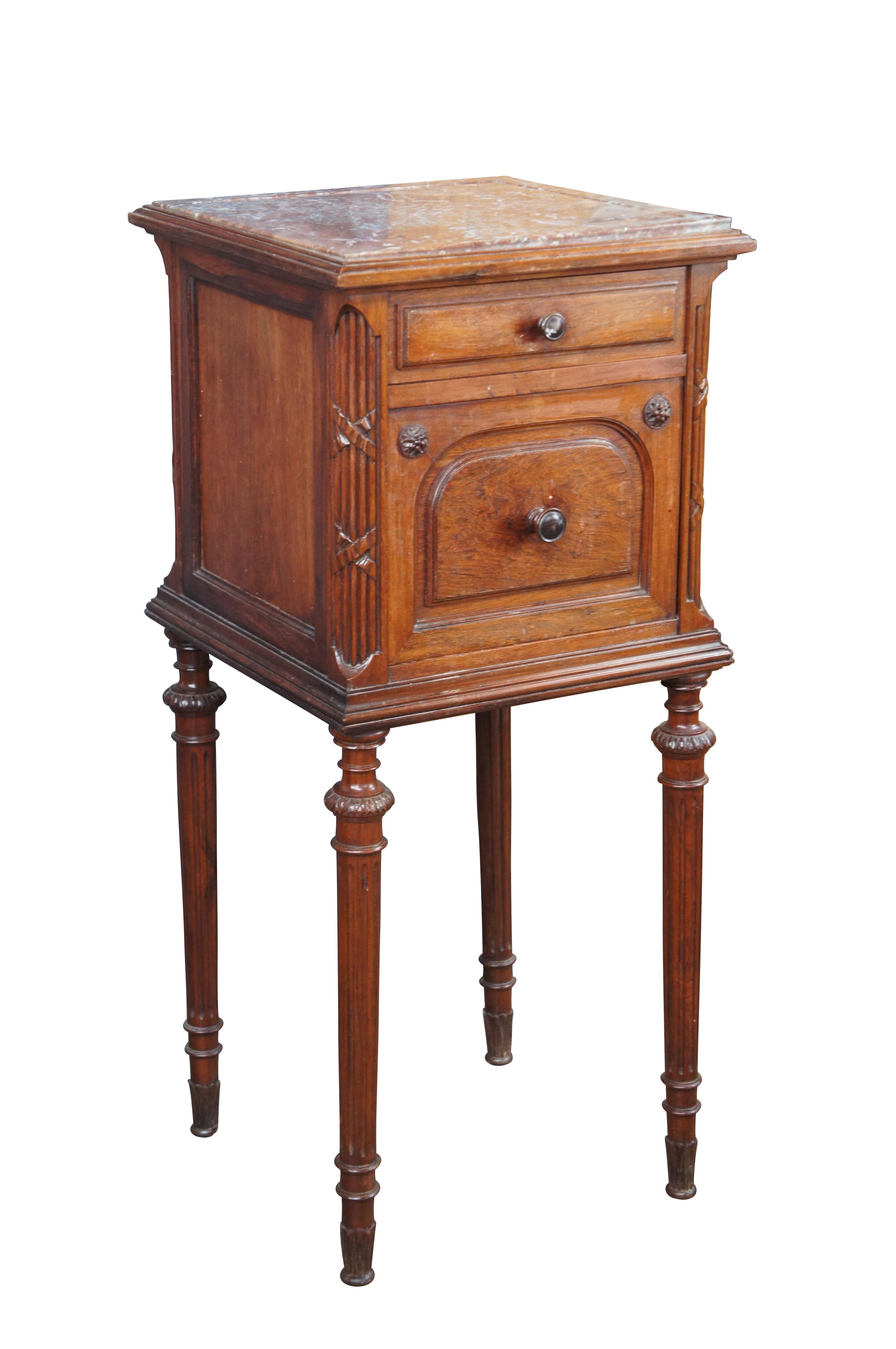 An antique late 19th Century French bedside table. Features a square frame made from walnut with inset rouge marble top. The case is accented by French medallions and ornate reeded and ribboned stiles. Includes one upper hand dovetailed drawer and a