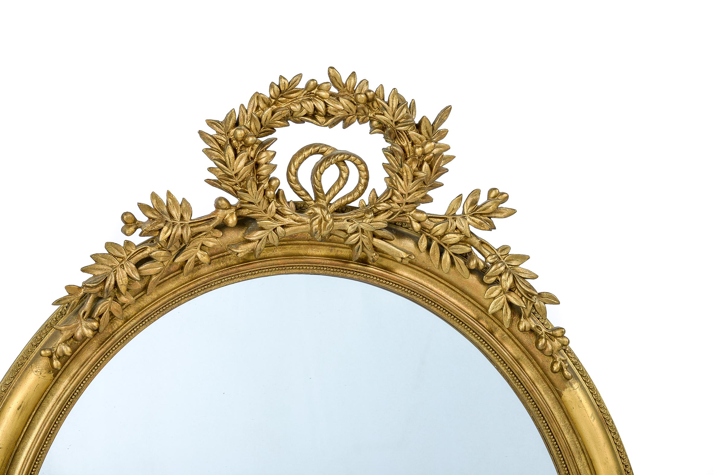 A beautiful antique French oval mirror that was made in France, circa 1880.
The mirror frame is decorated with an elegant top ornament. It features a berried laurel wreath tied together with a rope bow. Laurel has a very rich symbolic meaning in