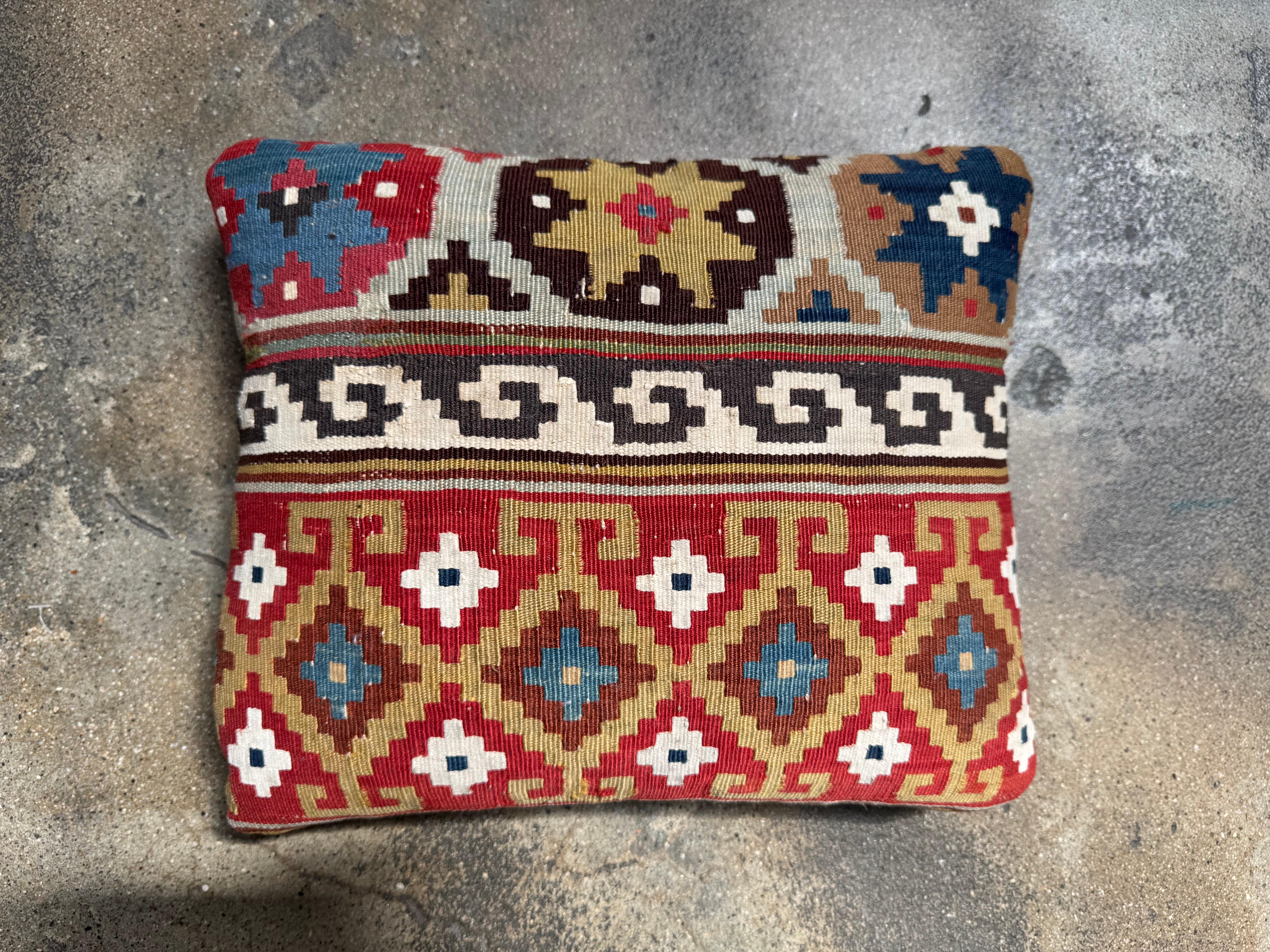 Ce coussin kilim caucasien antique du XIXe siècle offre un mélange vibrant de tradition et de polyvalence en matière de design. Il est tissé à la main à partir de laine richement teintée dans des tons cramoisis, ivoire, ocre, bleu profond et marron