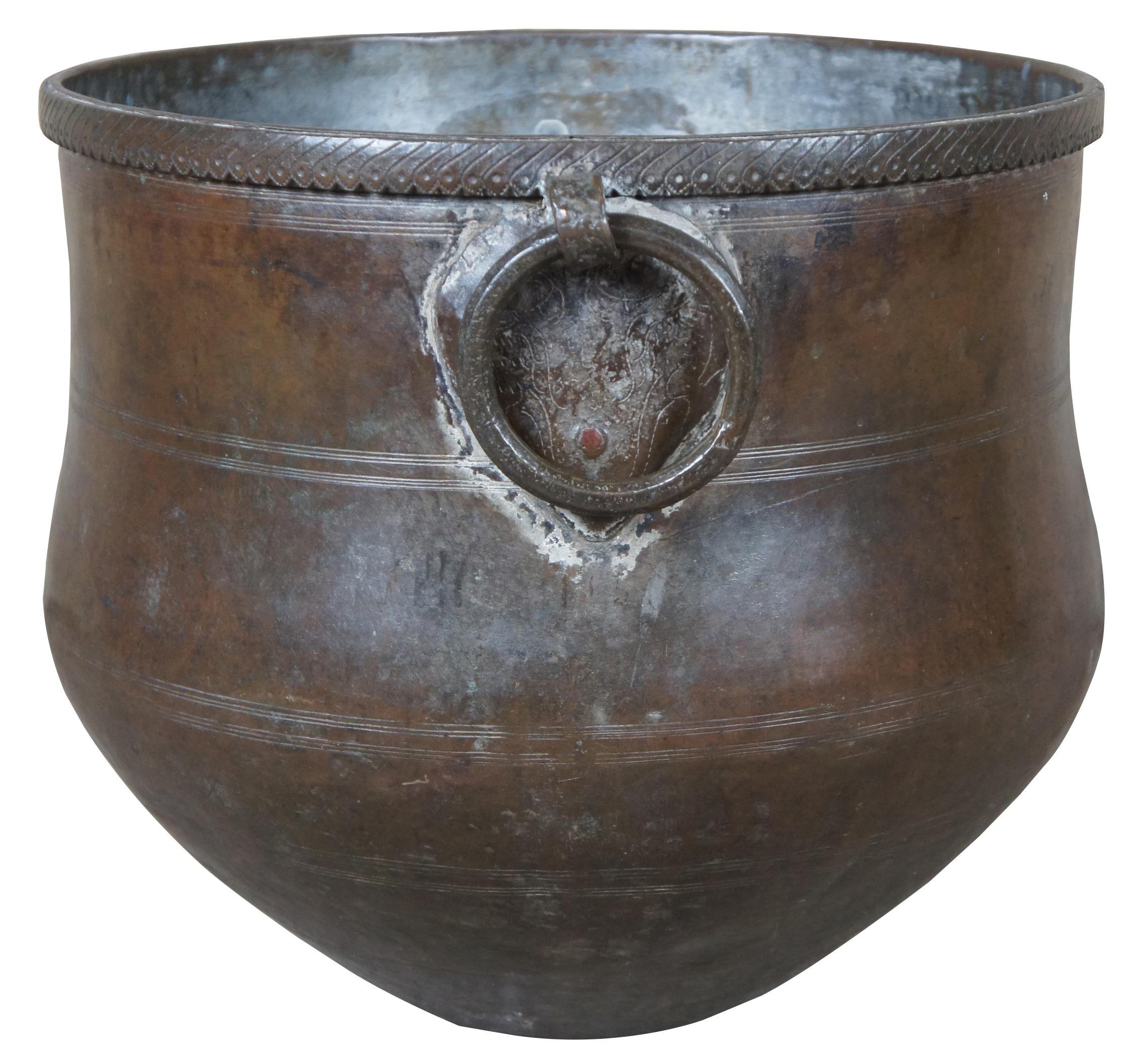 Un monumental récipient en cuivre martelé ou un pot de stockage d'eau. Rond avec une base effilée et des poignées en anneau de fer. Le bord est en fer ciselé. 
   
