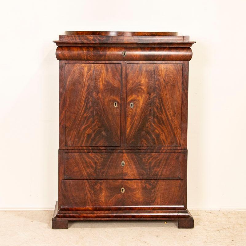 Antique flame mahogany 2 door cabinet from Denmark, circa 1860-1880. Made in two sections, there are 2 drawers below and 2 doors with a single drawer above. The entire front is covered in an exceptional veneer of book matched flame mahogany. This