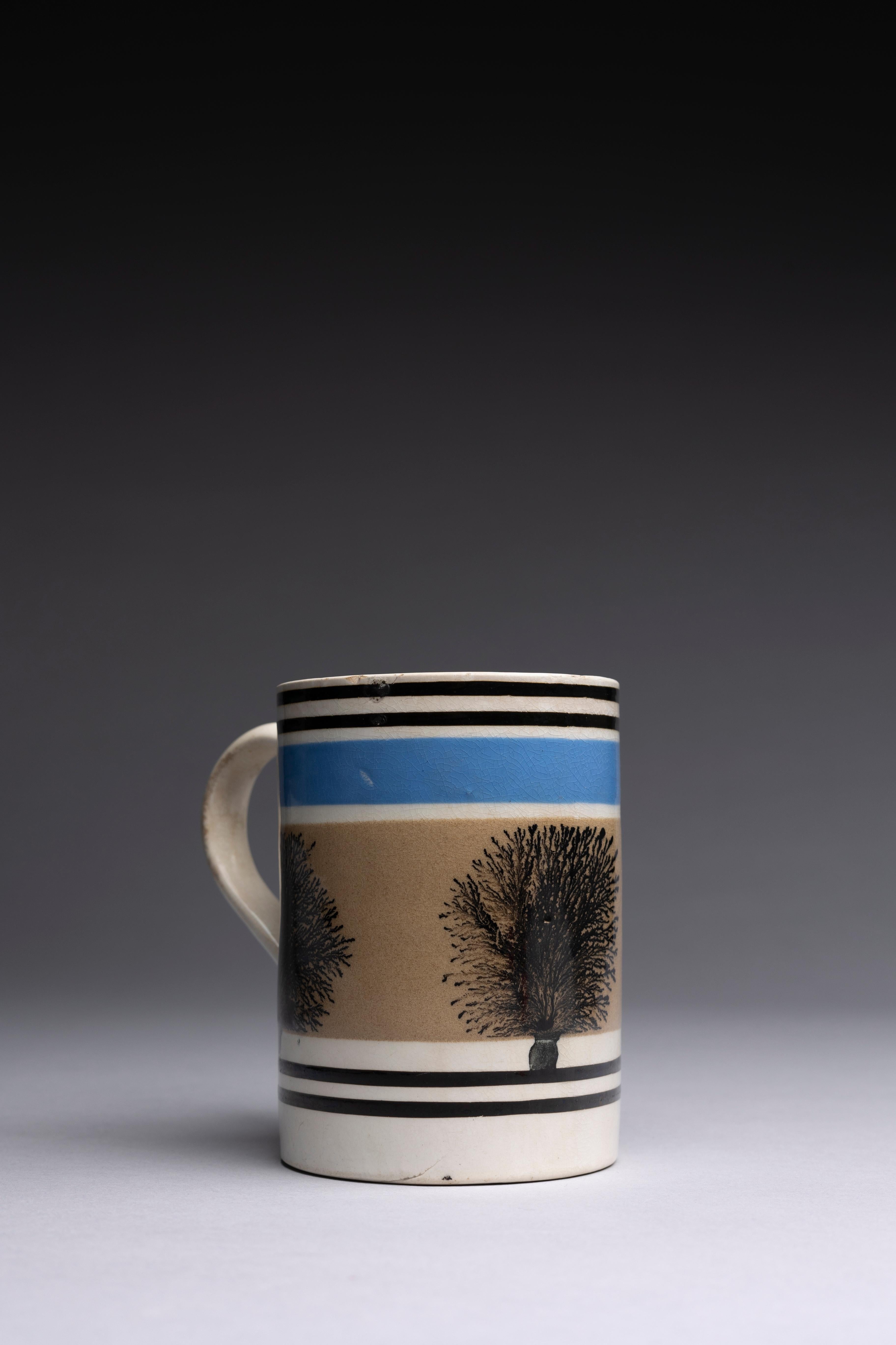Ein englischer Mochaware-Becher mit schwarzen und blauen maschinell gedrehten Bändern und einem Baummuster auf einer weichen braunen Glasur.

Dieser große Becher aus Mokkaware zeigt schwarze und blaue Rauten, die ein baumartiges Muster (Algen