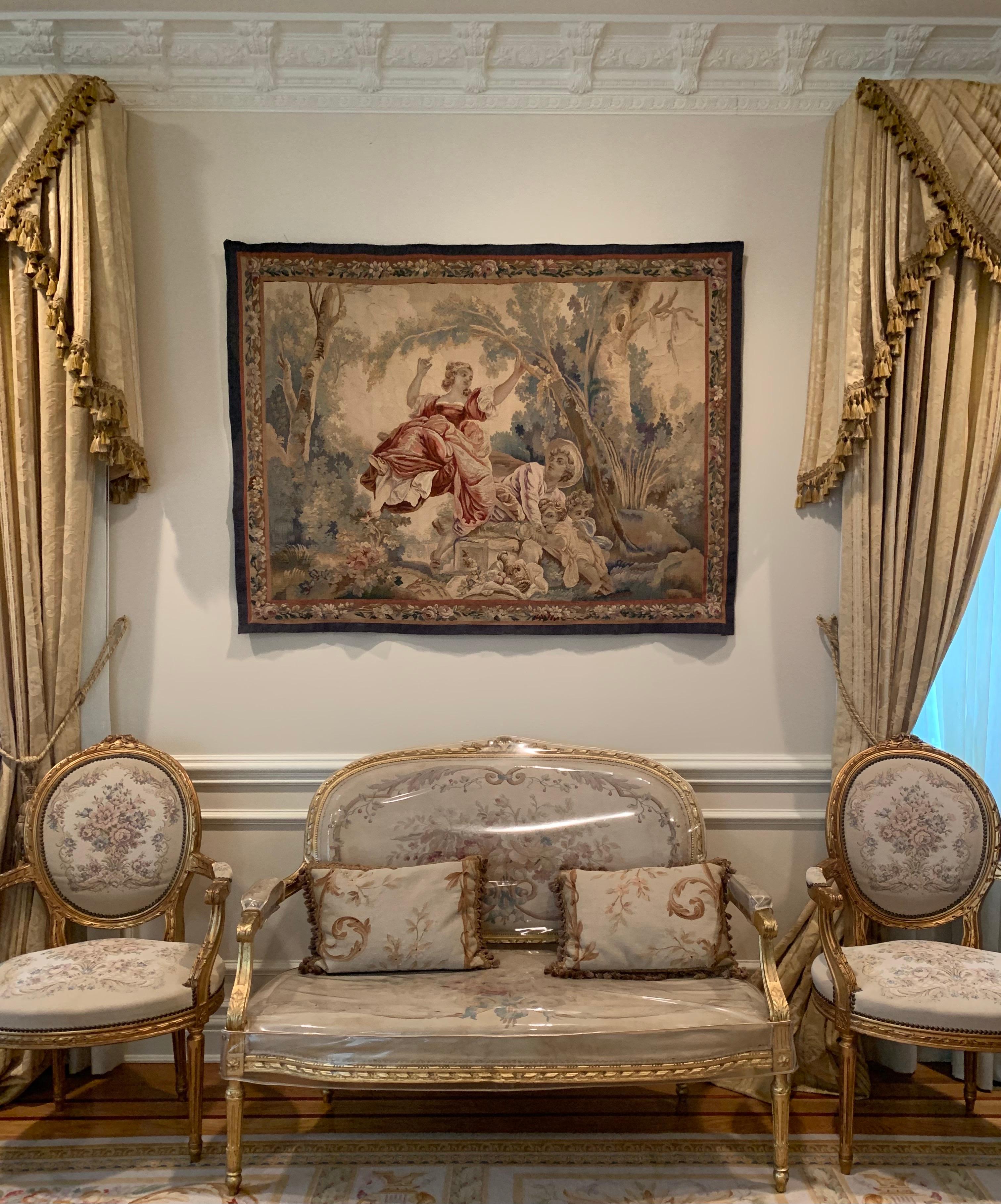 This is a lovely antique 19th century square French Aubusson tapestry depicting a on a beautiful spring day in the countryside with lush trees and vegetation with a woman on a swing and her husband and two children beside her. 

It is a lovely silk