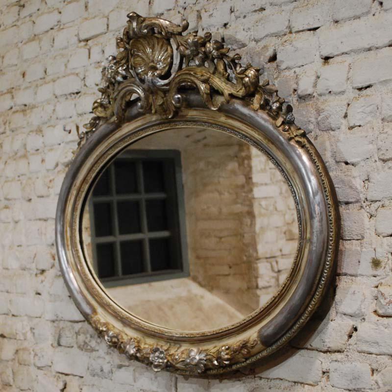 This beautiful oval mirror is partially silver leaf gilt. It has remnants of gold paint and it shows the bone color of the gesso.
It features a top cartouche with a central scallop flanked by C-scrolls, flowers, and leafs. This mirror was