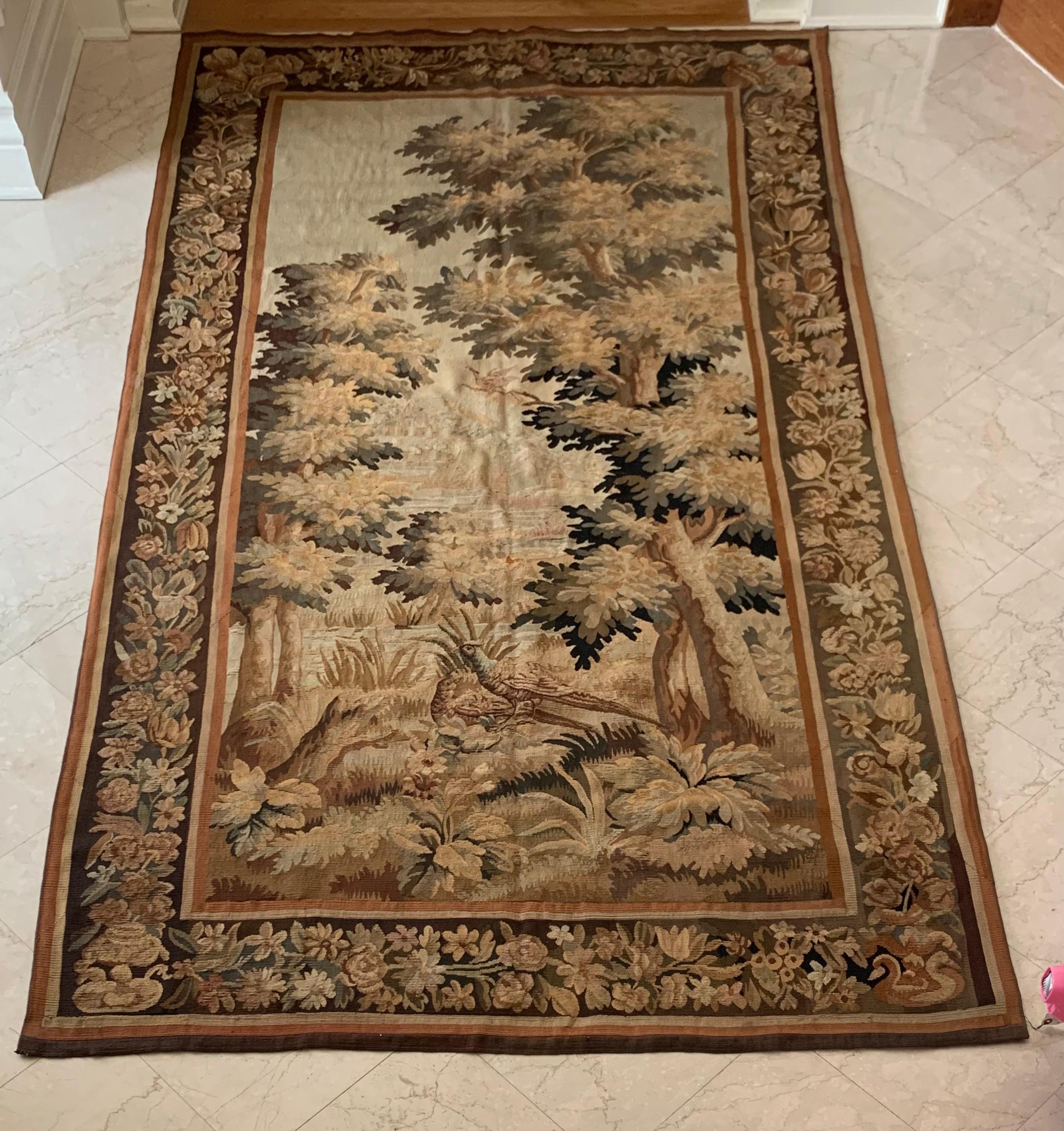 This is a lovely antique 19th century French Verdure Landscape Tapestry depicting a beautiful summer scene of a countryside with lush trees and birds. It has an ornate border of flowers and garlands and a classic example of French Verdure tapestries