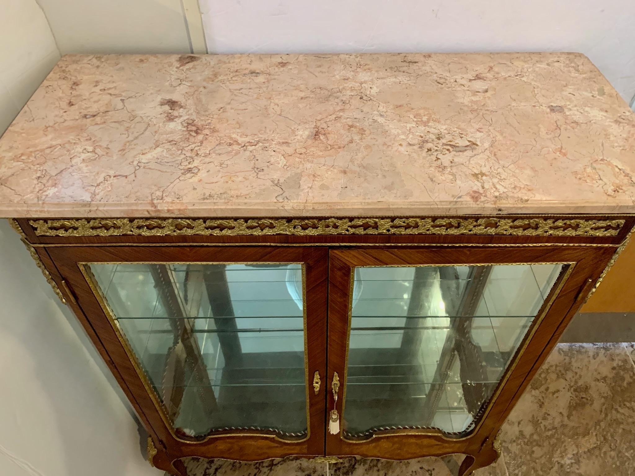 Exquisite French vitrine has Louis XVI styling with bronze ormolu mounts and marble top. Twodoors and both sides have glass. The interior has glass shelves in front of a mirrored back.